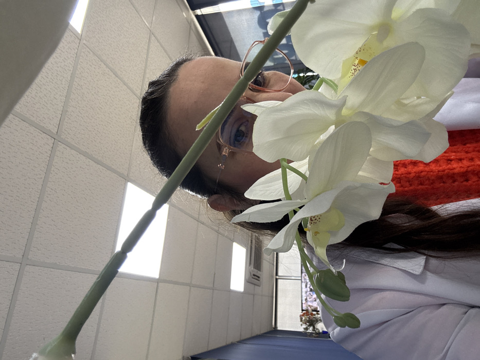 The image depicts an indoor scene in the city of Meknes, Morocco. The primary subject is a female individual, partially visible behind a foreground element. Her upper face, including her eyes and forehead, is visible. She wears eyeglasses with rose-gold frames and her dark hair is pulled back. She is dressed in a white lab coat or similar professional attire over a bright red garment, possibly knitted. Prominently in the foreground, partially obscuring the subject, is a branch of artificial white orchid flowers with yellow centers and several green buds. The green stem of the orchid extends diagonally across the upper portion of the frame. The background features a drop ceiling with white square acoustic tiles and recessed rectangular fluorescent light fixtures. To the left, a light blue wall section is visible, along with an air conditioning unit and a distant window with horizontal blinds. The subject's gaze is directed towards the camera.