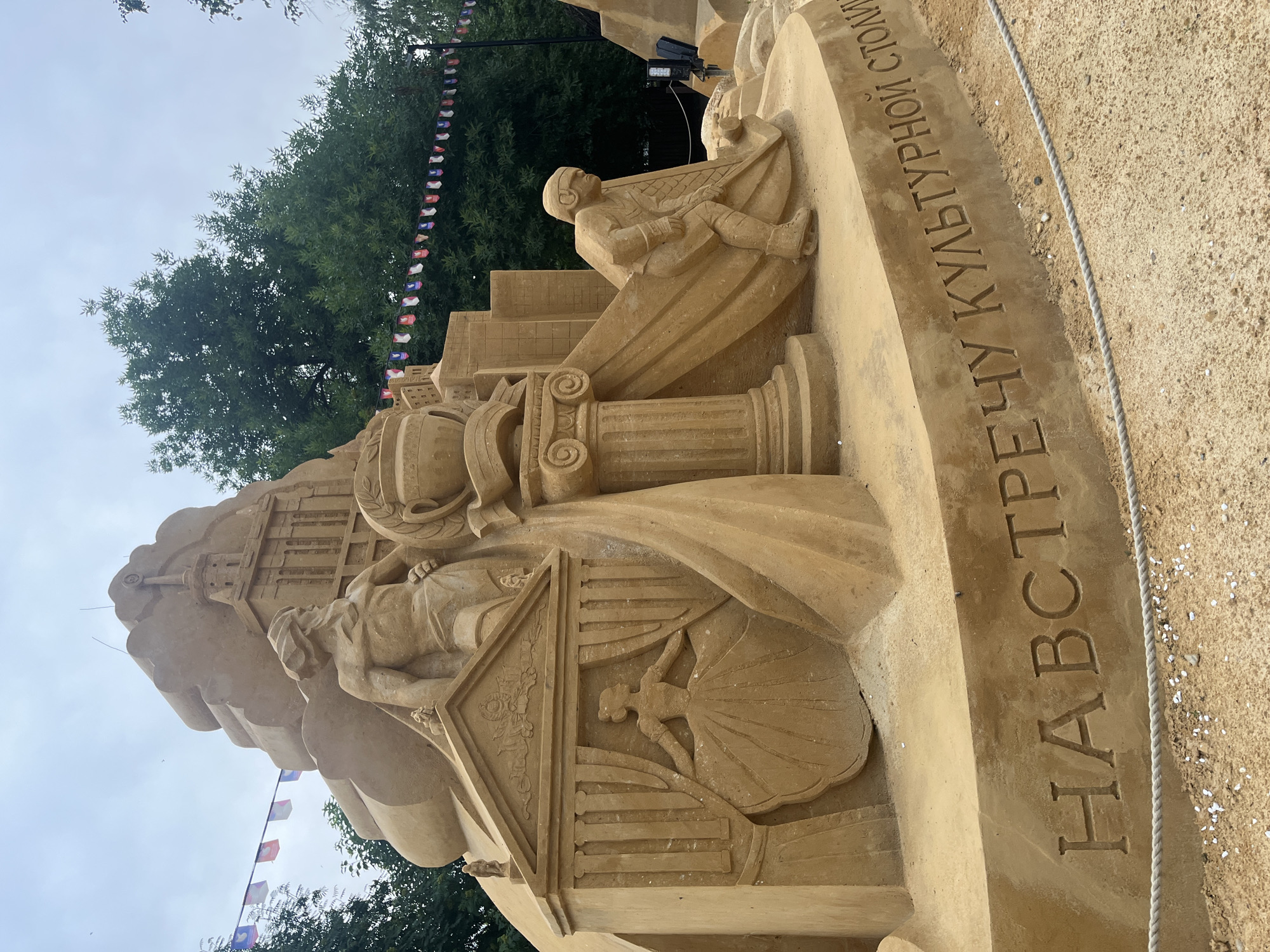 This media file showcases a large, intricate sand sculpture, likely an exhibit from a festival or event. The sculpture is primarily made of sand and depicts a complex scene with human figures and architectural elements.
The main subjects include at least two human figures rendered in sand. One figure appears to be seated, possibly playing an instrument or holding something, and is wearing a helmet and clothing reminiscent of an aviator or historical figure. Another figure is depicted in a more stylized, almost bas-relief form within an architectural detail, appearing to be a dancer or performer in a flowing gown. Architectural elements such as columns with Ionic volutes and layered structures resembling buildings are incorporated into the sculpture, suggesting a grand or historical theme.
The setting appears to be outdoors, with trees visible in the background and a cloudy sky overhead, suggesting it might be daytime. Small flags, possibly representing different nations or organizations, are strung up behind the sculpture, hinting at an international event or exhibition. A rough rope barrier is visible in the foreground, indicating the sculpture is on display and protected from direct public access.
A prominent detail is the carved text on a curved surface of the sculpture. The text is in Cyrillic script and appears to read "НАВСТРЕЧУ КУЛЬТУРНОЙ СТОЛИЦЫ" which translates to "Towards the cultural capital." This text provides a significant clue to the theme or purpose of the sculpture, likely celebrating or anticipating a city being designated a cultural capital. The sand itself is a warm, light brown color. The overall impression is one of artistry and community engagement, with the sculpture serving as a focal point for a public event.