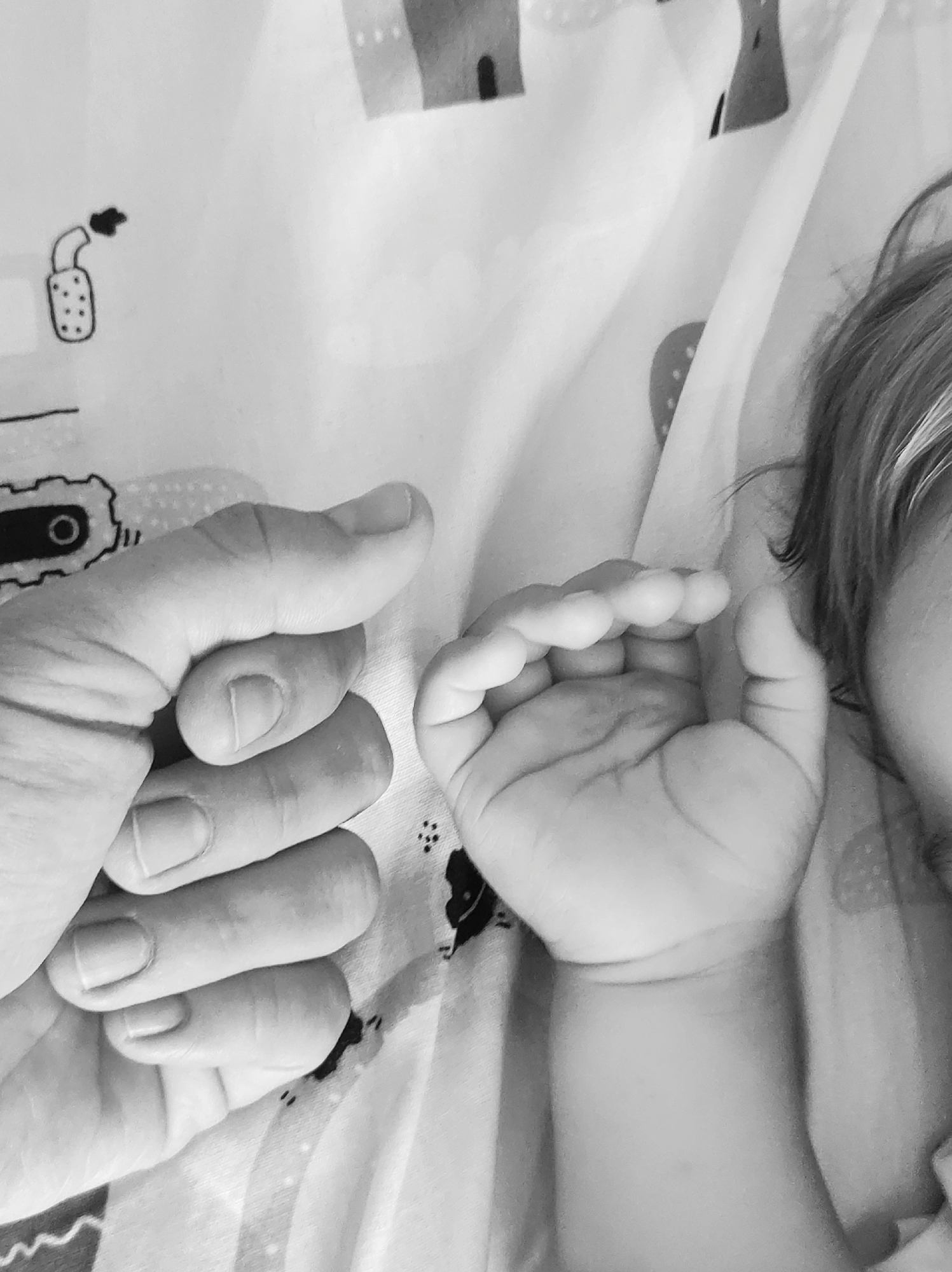 The image shows a close-up of an adult's hand holding a baby's hand. The background is a white fabric with a repeating pattern of black drawings of houses and other objects. The image is in black and white. There is no information about the location in the city of Chaikovsky, Russia.