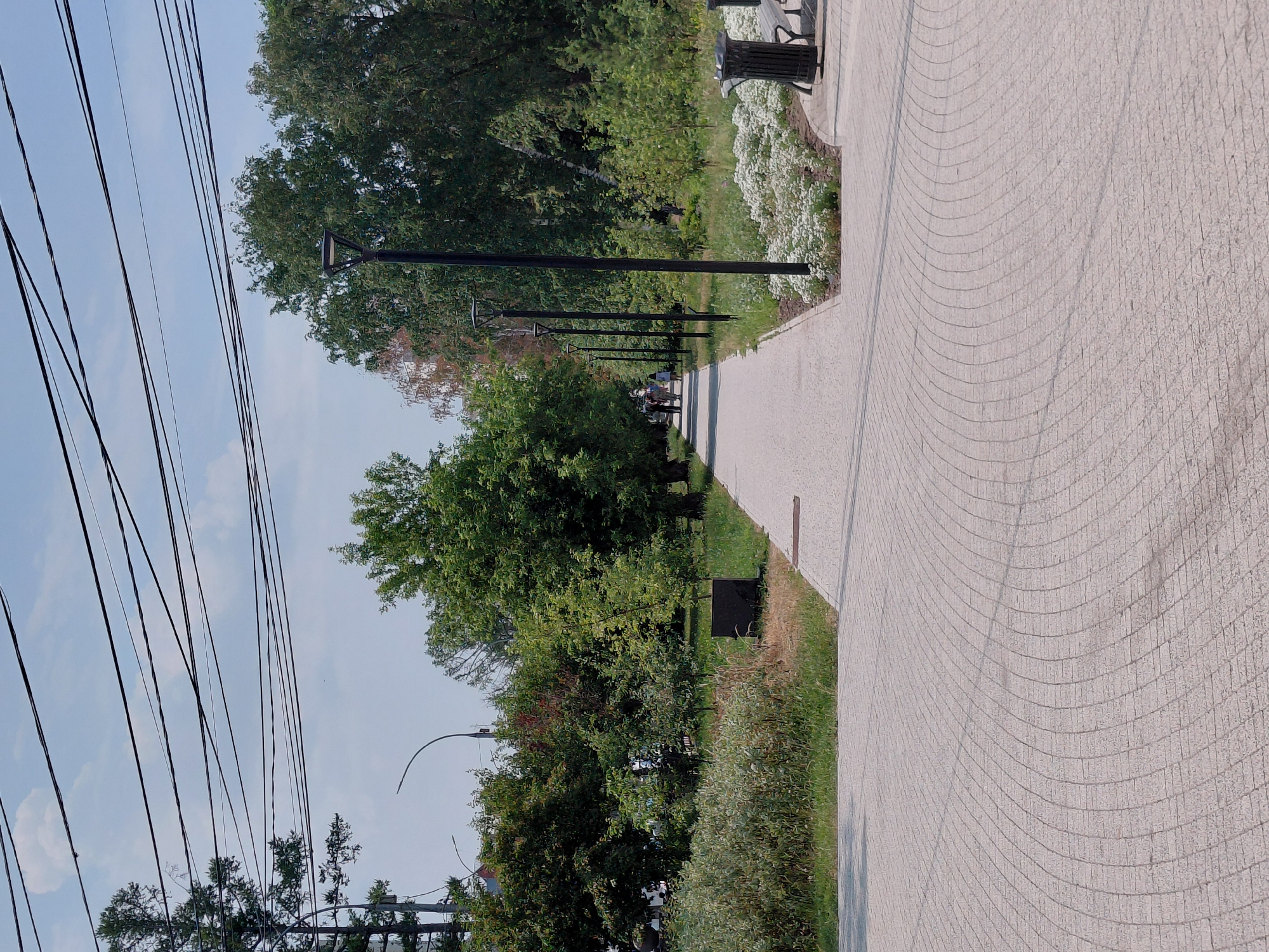 The image depicts a paved pathway in a park setting, likely in the city of Novosibirsk, Russia, given the common architectural and landscaping style. The pathway is constructed of light-colored paving stones laid in a curved pattern, leading into the distance. It is bordered by green grass and lush trees on both sides. In the foreground, the pathway curves to the right, revealing more of the paving stone pattern. Several modern lampposts are visible along the pathway, suggesting it's a public space designed for evening use. In the background, a few people are walking along the path, their forms somewhat indistinct due to the distance. The sky above is a pale blue with scattered white clouds. A prominent feature is the network of overhead power lines, a common sight in urban environments. Further along the path, to the right, there is a park bench and a trash receptacle. The overall scene conveys a sense of a well-maintained urban park, inviting for leisure and recreation.