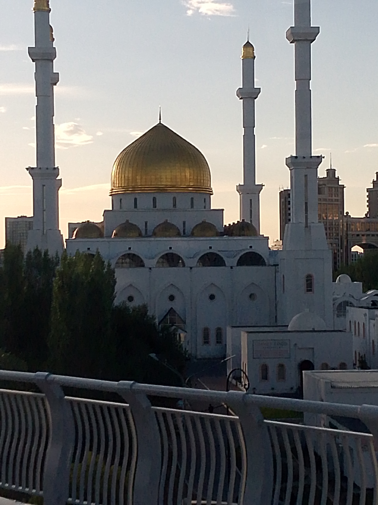This media file features a view of the Hazrat Sultan Mosque in Astana, Kazakhstan. The mosque is a prominent landmark, characterized by its large golden dome and multiple white minarets. The image captures the mosque's white facade with its distinctive arched windows and smaller golden domes. In the foreground, a modern metal railing of a bridge or walkway is visible, suggesting the viewpoint is elevated and some distance from the mosque itself. Lush green trees partially obscure the lower sections of the mosque, adding a natural element to the urban landscape. In the background, contemporary high-rise buildings rise above the trees, indicating a modern city setting. The sky is a soft, pale blue with scattered clouds, and the warm, golden light suggests it is either early morning or late afternoon, possibly during sunset. No people are visible in the frame. There is no visible text.