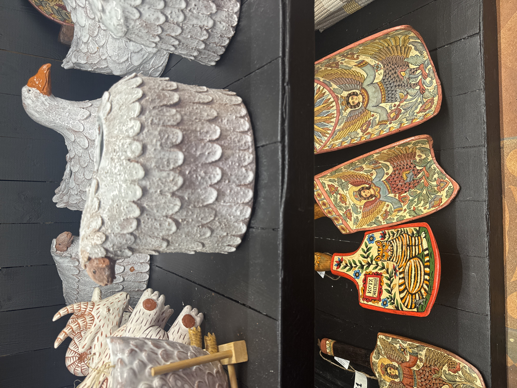 The image displays a close-up view of decorative items on two black wooden shelves. On the top shelf, several ceramic animal figures are visible, including sheep with textured bodies and heads, and white geese with orange beaks. Some of these appear to be planters or vases. A goat figure with horns and patterned markings is also present, along with other indistinct ceramic objects.
The bottom shelf features three painted wooden decorative plaques. The leftmost plaque depicts a winged figure with outstretched arms, rendered in a folk art style. The central plaque is shaped like a cutting board and prominently features a painted orange and black striped cat sitting in front of a floral design, with the Russian text "КОТ ЗНАЙКА" (Kot Znaika, meaning "Know-it-all Cat") in a decorative frame. To the right of the cat plaque is another plaque similar to the leftmost one, also depicting a winged figure with outstretched arms and ornate floral motifs. The plaques are arranged diagonally on the shelf.
The overall aesthetic suggests a display of traditional Russian folk art, likely found in a shop or market. This scene is consistent with the type of handicrafts sold in Suzdal, Russia, a city renowned for its well-preserved historical architecture and vibrant folk art traditions.