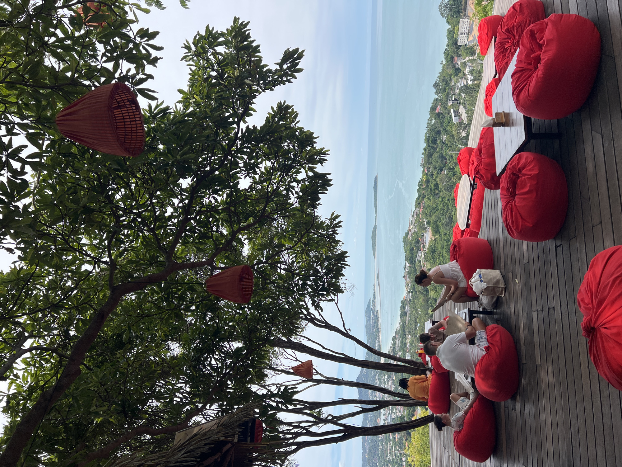In Baan Chaweng Noi, Thailand, a scenic open-air restaurant offers a panoramic view of the turquoise sea and a lush green hillside. The foreground is dominated by a wooden deck furnished with numerous vibrant red beanbag chairs and low wooden tables. Several groups of people are seated, some engaged in conversation, others reading, while enjoying the stunning vista. A few red, conical lamps hang from the branches of large trees that provide ample shade over the seating area. The background reveals the coastline dotted with buildings and the vast expanse of the ocean meeting the horizon under a slightly overcast sky.
