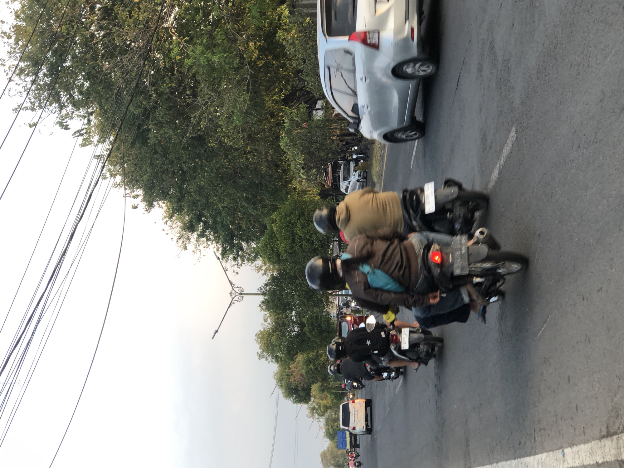 The image depicts a street scene in Siring, Indonesia, during daylight. Several motorcycles are in motion on a paved road. In the foreground, two individuals on a motorcycle are visible from behind, both wearing helmets. The passenger has a turquoise bag slung across their chest. To their left, another motorcycle with two riders, also wearing helmets, is slightly ahead. Further down the road, more motorcycles and a white car are visible. A silver minivan is in the right lane, traveling in the same direction. The environment includes trees lining the road and overhead power lines. The sky is overcast, suggesting it may be late afternoon.