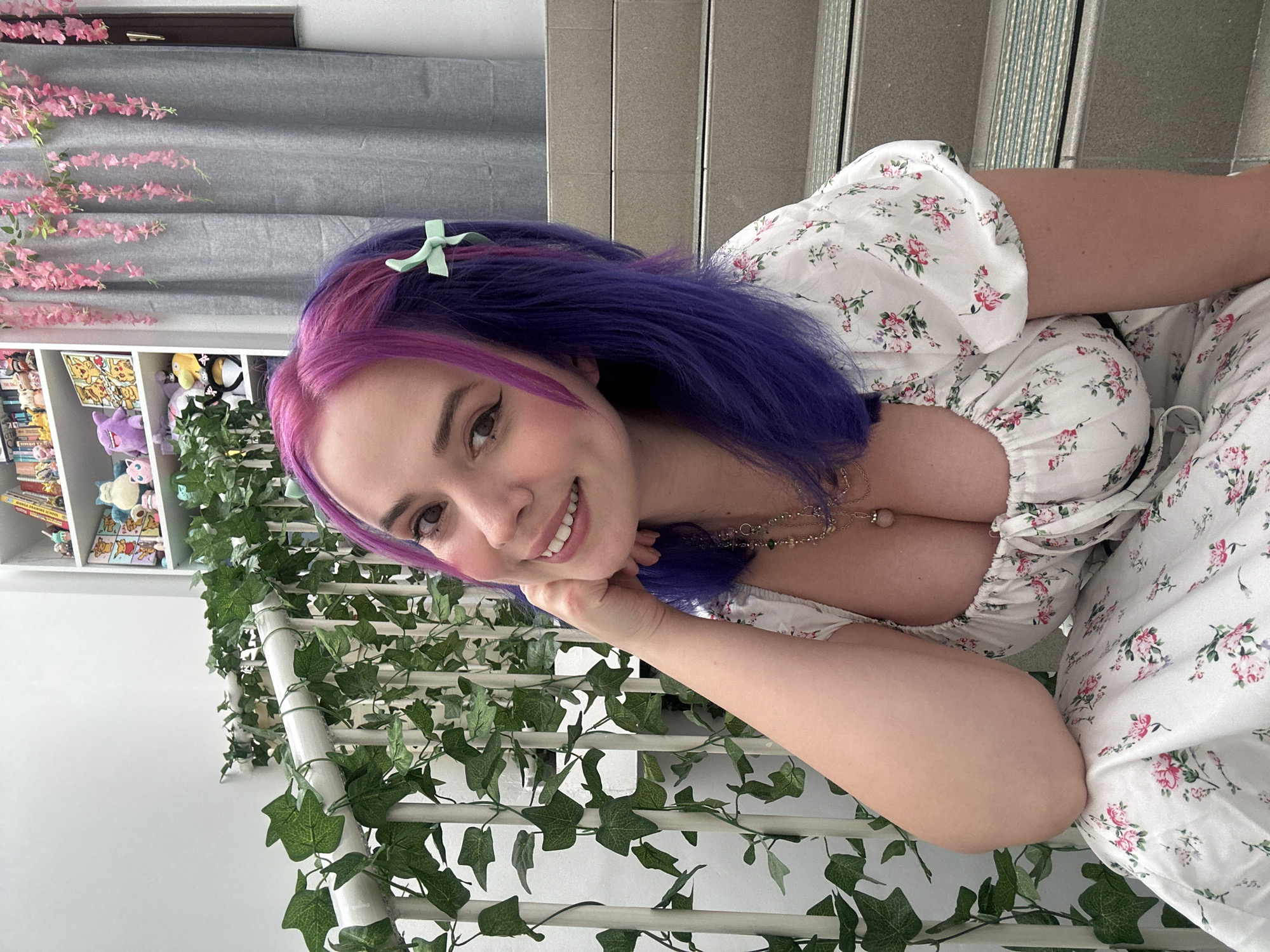 A person with pink and purple hair is smiling at the camera. They are wearing a white floral print dress and leaning against a white railing covered in fake ivy. Behind them is a bookshelf filled with various items and a gray curtain. In the background, there are stairs with silver accents and a gray wall. The location is possibly indoor, somewhere in the city of Zagreb, Croatia.