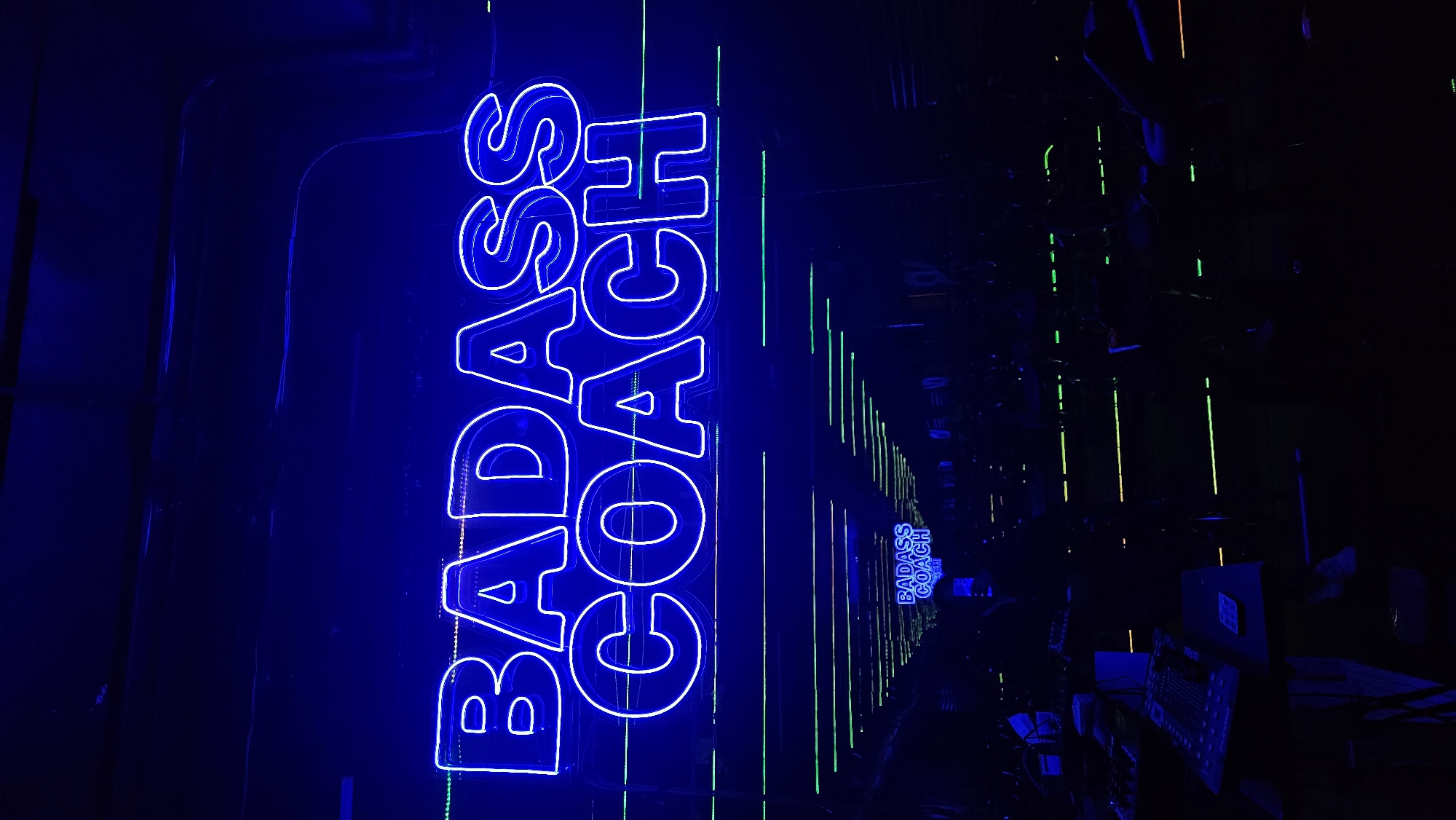 The image shows a neon sign that says "BADASS COACH" in a vertical format, illuminated in bright blue. The sign is positioned against a dark background with a reflective surface. The reflections create an illusion of depth, with multiple smaller versions of the sign receding into the distance. Green and yellow vertical lines, also reflections, add to the visual effect. The location is in the city of Puebla, Mexico. The reflections suggest a mirrored environment, possibly in an indoor setting. The image contains objects like desks, keyboards, and screens.