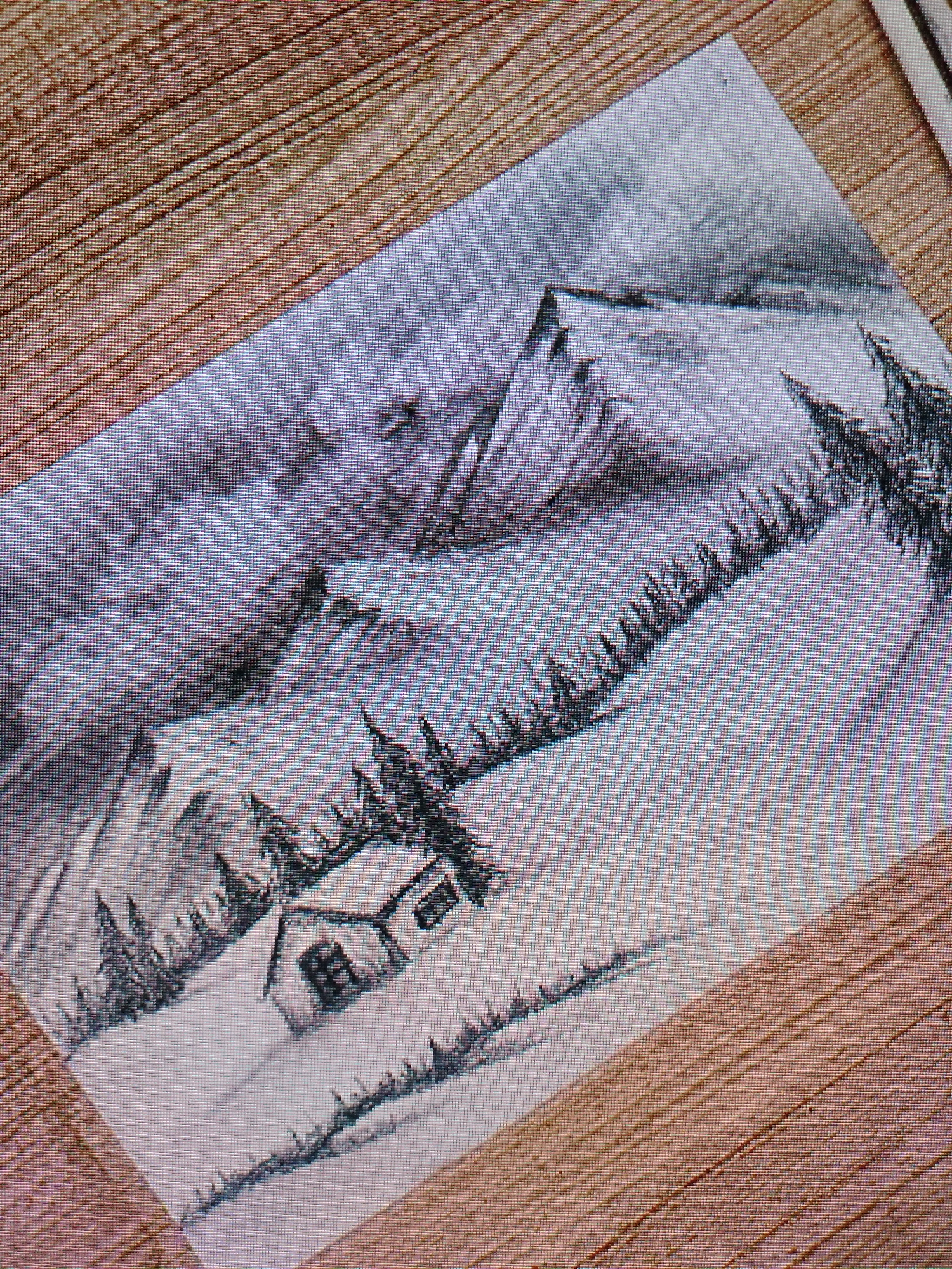 The image shows a drawing of a snowy mountain landscape. In the foreground, there is a small cabin nestled in the snow, surrounded by pine trees. Behind the cabin, a snow-covered mountain rises, with a line of trees extending up the slope. Further back, additional mountains are visible under a cloudy sky. The drawing is placed on a wood-grain surface.