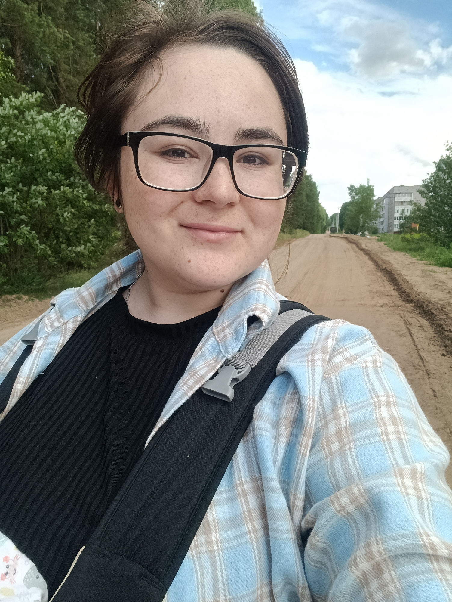 A young person with glasses and brown hair takes a selfie outdoors. They are wearing a black ribbed shirt and a blue and white plaid shirt. A black backpack strap is visible. The background shows a dirt road lined with trees and buildings, suggesting an urban or suburban environment. The sky is partly cloudy. The location is the city of Noviy, Russia.