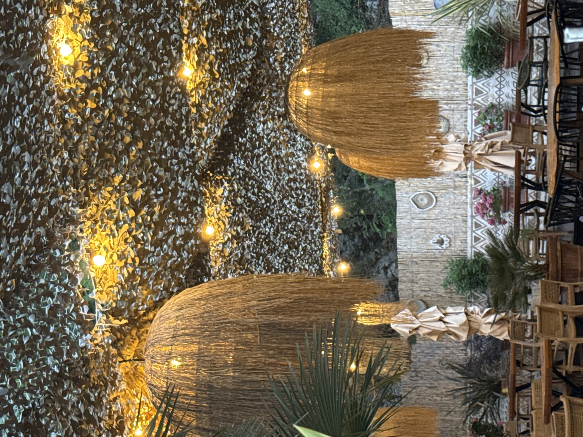 This media file captures an outdoor setting with a focus on decorative elements and dining furniture. The scene appears to be set at dusk or evening, as evidenced by the string lights illuminating the area. The background features a textured wall covered in what looks like foliage or a patterned textile, interspersed with glowing yellow lights.
Prominently featured are two large, dome-shaped objects made of straw-like material, possibly lampshades or decorative features, each with internal lighting casting a warm glow. Below these structures, draped fabric adds to the ambiance. In the background, a rustic wall with geometric patterns is visible, along with potted plants and greenery, suggesting an outdoor dining or lounge area.
To the right of the frame, a collection of wooden tables and chairs, some with woven seats, are arranged. This arrangement indicates a dining or seating area. The overall impression is one of a relaxed, perhaps restaurant or cafe setting, in Kamenovo, Montenegro, designed to create a cozy and inviting atmosphere. There is no visible text in the image.