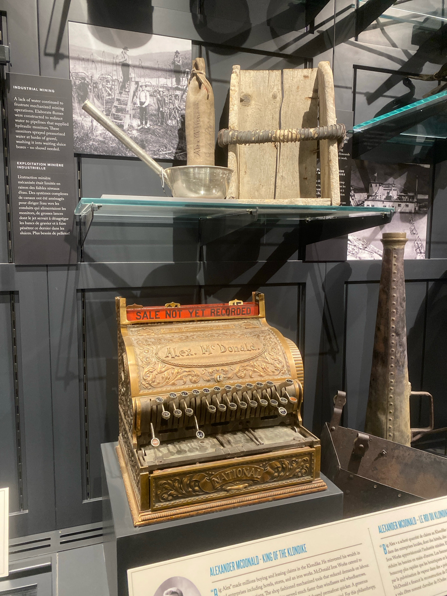 This image depicts a museum exhibit in Dawson City, Canada, focusing on Klondike gold rush history. In the foreground, a vintage National cash register, ornately decorated and labeled "Alex. McDonald.", sits on a black pedestal. To its right, a weathered metal scoop or trough is visible. Behind the cash register, on a glass shelf, are artifacts related to industrial mining. These include a large silver pot with a long handle, a wooden bucket with a metal band, and a canvas-wrapped object with writing. Above these, a black and white photograph shows several men working with equipment, likely related to mining operations. A sign to the left of the photograph, titled "INDUSTRIAL MINING," provides historical context. Further to the right, a tall, conical metal object with rivets, resembling a megaphone or horn, stands upright. The overall environment suggests a well-lit museum display with dark gray paneling on the walls.
