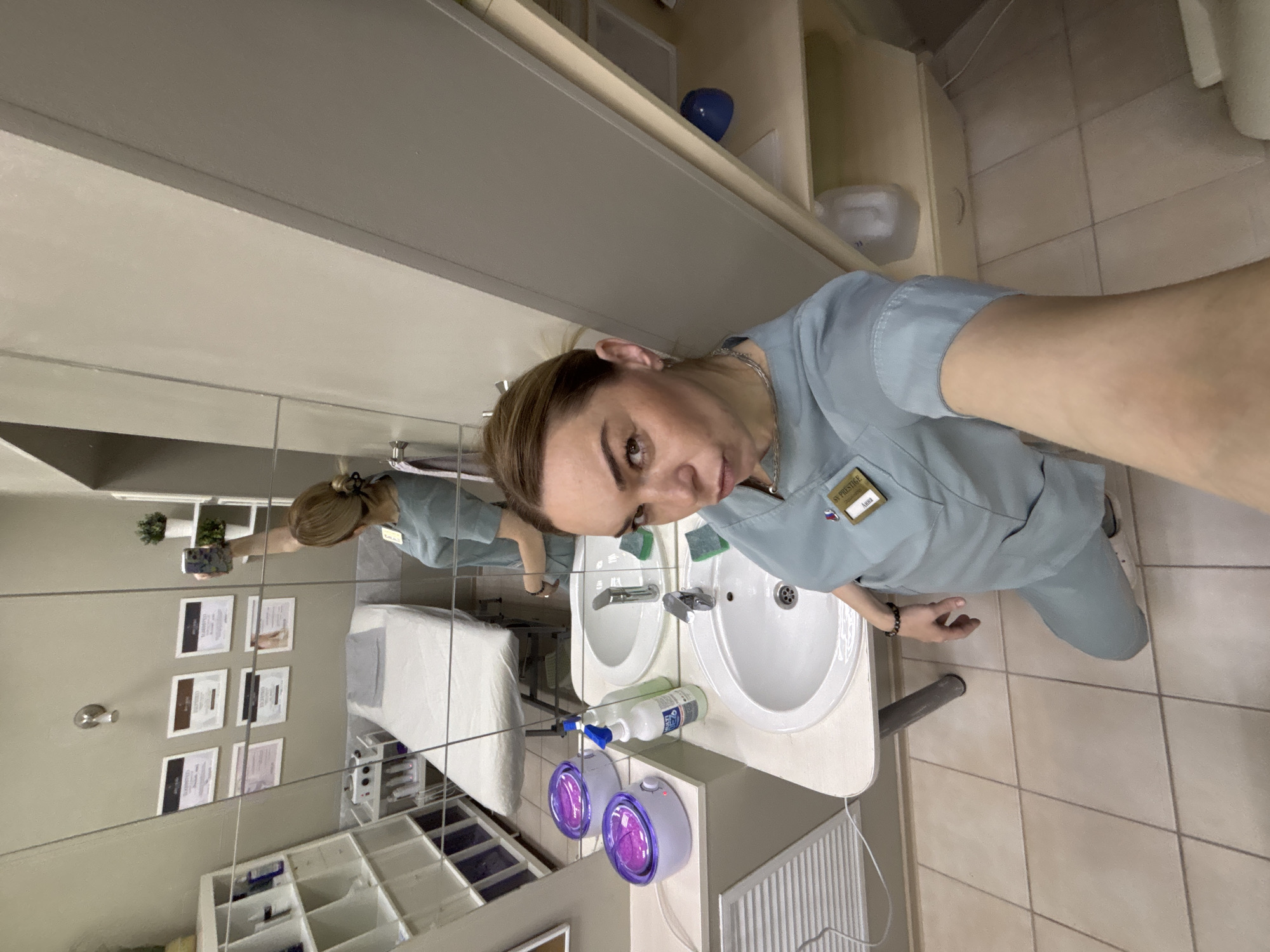The image is an angled, indoor shot of a woman in a light blue uniform standing in front of a mirror in what appears to be a professional setting, possibly a salon or medical spa. The reflection in the mirror shows a treatment bed, shelves with supplies, and framed certificates on the wall, suggesting a place of business. The woman is holding a phone, possibly taking a selfie. The uniform has a name tag or badge labeled "SV PRESTIGE Anna". The location is Sinyavino, Russia.
