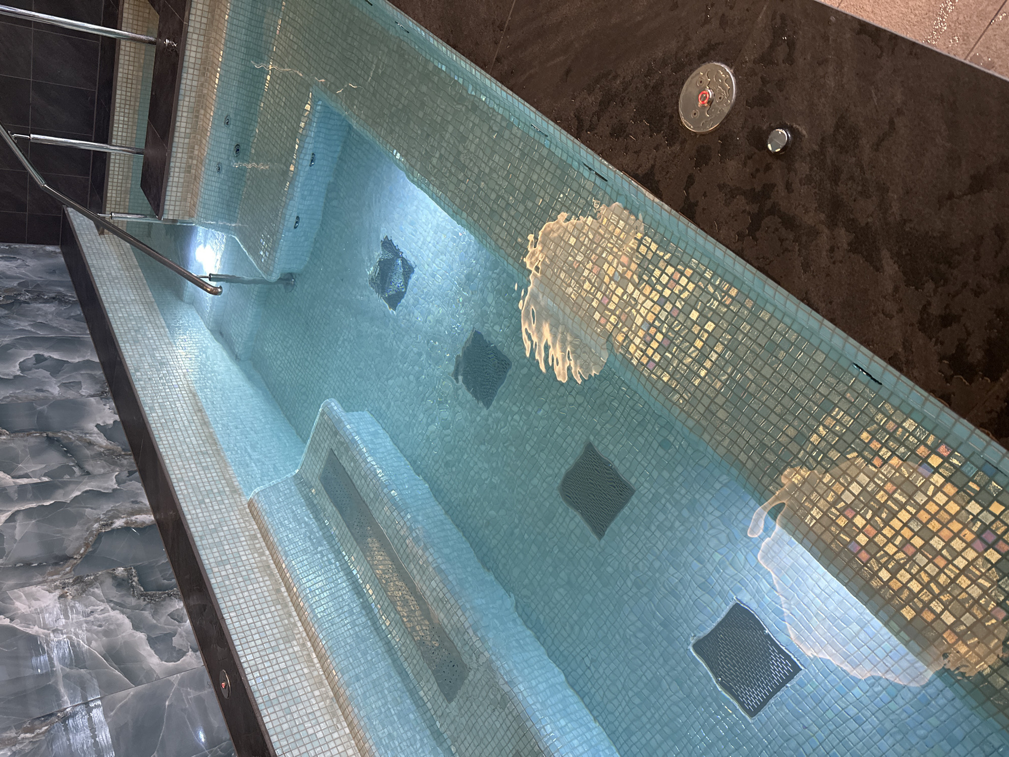 The image displays an indoor swimming pool or hot tub, viewed from an elevated, oblique perspective. The rectangular pool is meticulously lined with small, iridescent light blue and aqua mosaic tiles, which shimmer under the water. Integrated steps, also tiled with the same mosaic, are visible at the left end, accompanied by a sleek, polished metal handrail for entry and exit.
The clear water in the pool reflects ambient light, creating dynamic, undulating patterns across its surface, especially prominent on the far tiled wall and near the bottom. Several dark, square-shaped grates are visible on the pool's floor, suggesting water circulation or jet features.
The immediate decking around the pool is surfaced with dark, possibly black or very dark grey, textured tiles that appear wet in places. On this dark surface, two circular metal fixtures are positioned: a larger one with a distinctive red button or indicator, and a smaller, plain silver button. These are likely controls for the pool's functions, such as jets, lighting, or temperature.
To the left of the pool, the flooring transitions to an elegant, lighter grey tile with a swirling, marbled pattern. The overall scene suggests a modern and well-appointed spa, hotel, or private wellness facility. No people are present, indicating a tranquil setting ready for use. There is no visible text in the image. The setting is located in London, United Kingdom.