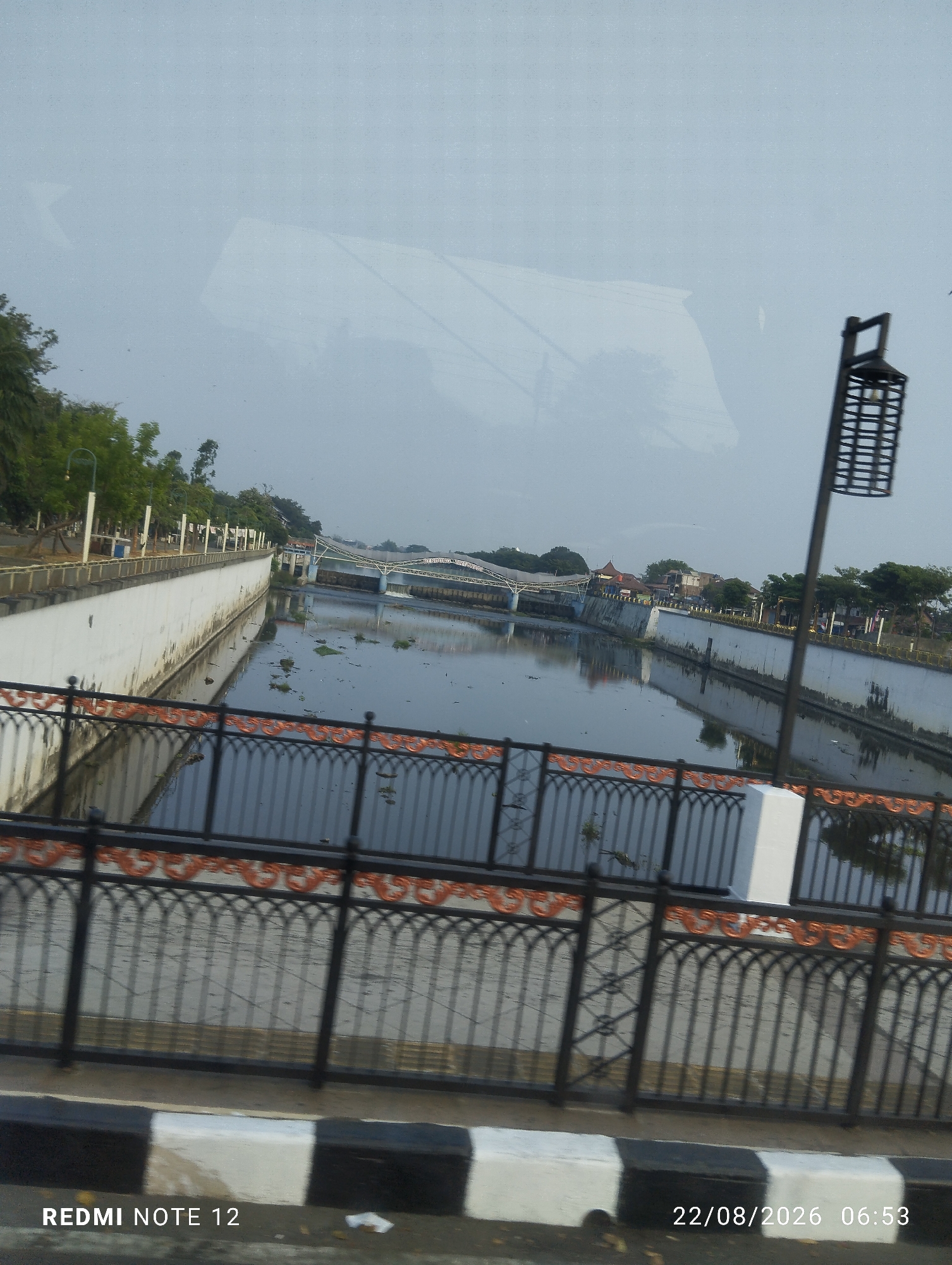 The image shows a canal in the city of Surakarta, Indonesia. The canal is lined with white walls and a black metal fence with ornate copper-colored accents. Trees and buildings are visible on the far side of the canal, and a bridge spans it in the distance. The water in the canal is calm and reflects the sky and surrounding structures. The overall scene suggests a public space within the city. A street curb with black and white stripes is visible in the foreground, along with a timestamp indicating the date and time the photo was taken.