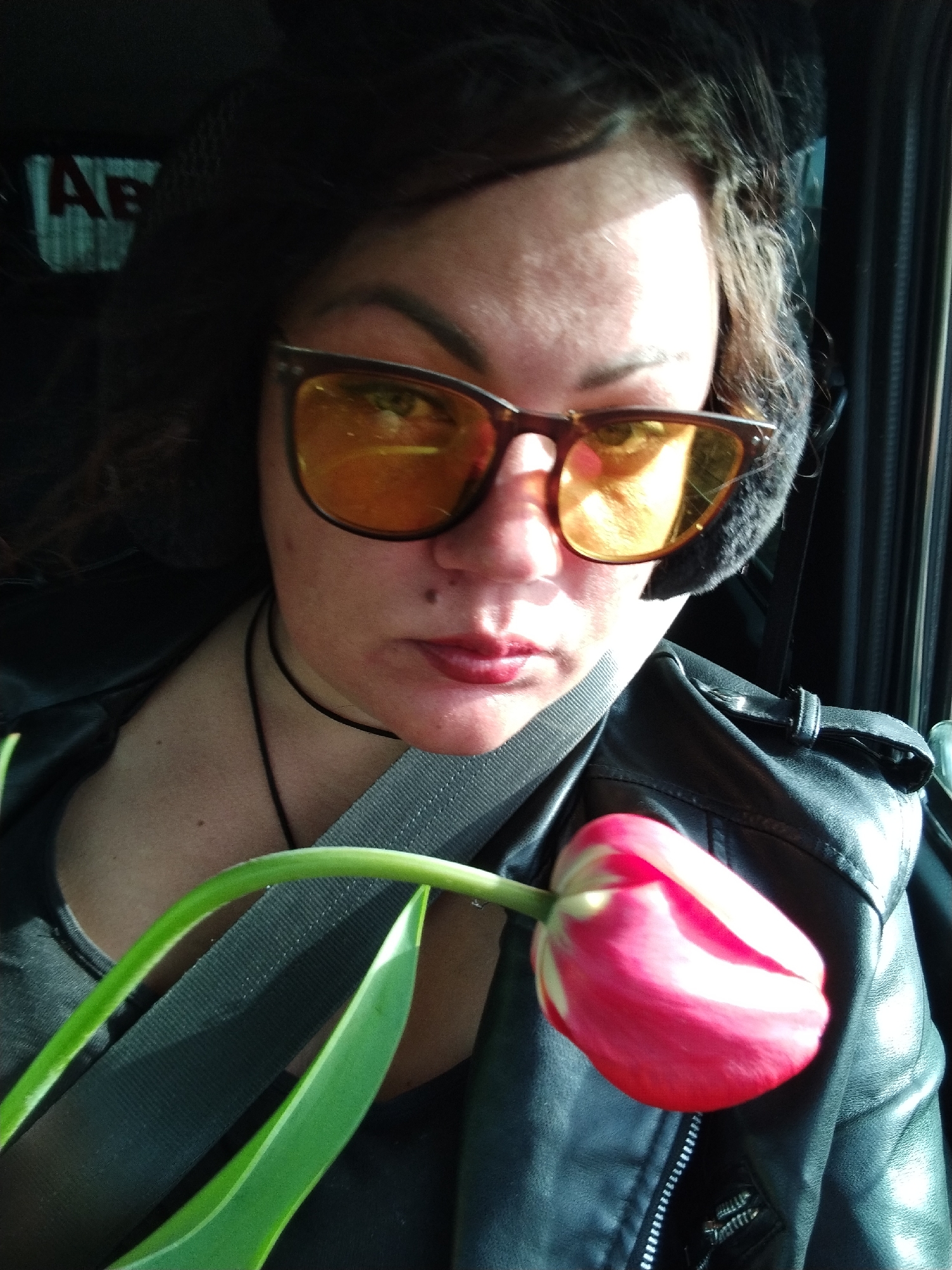 A woman is in a car wearing a black leather jacket, black shirt, seatbelt, a black choker necklace, and brown-framed sunglasses with yellow lenses. She also appears to be wearing furry ear muffs. She is holding a red and white tulip. The background shows the interior of a vehicle, with a glimpse of the outside through the window. The partial sign "AE" in the background suggests that the location is Chaikovsky, Russia.
