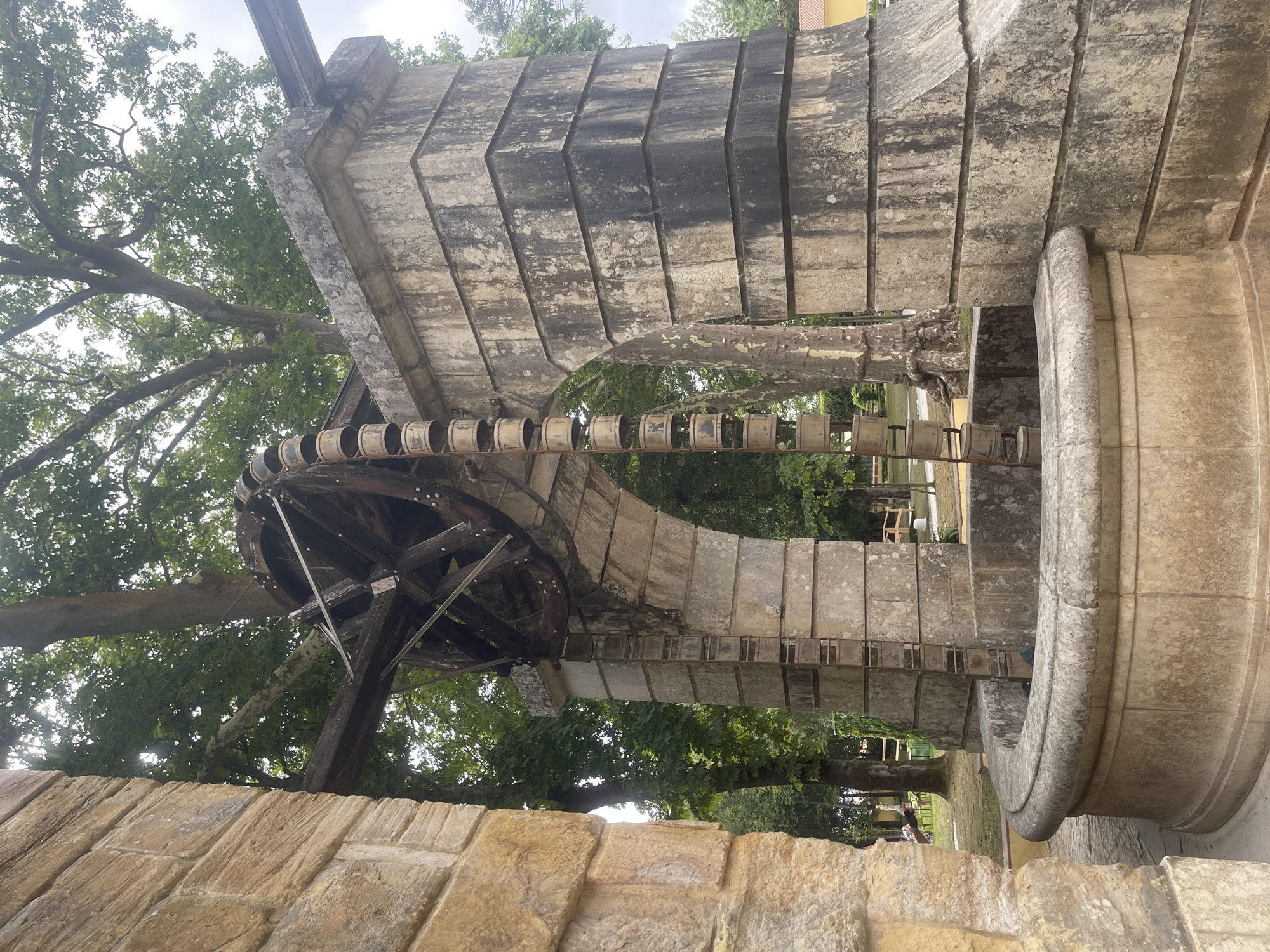 The image shows a weathered stone fountain with a large wooden wheel and a bucket chain mechanism, likely a water pump, set against a backdrop of trees. The stone structure exhibits signs of age and wear, with patches of dark discoloration. The fountain basin is circular with a thick, rounded rim. The bucket chain hangs vertically from the wheel, with many cylindrical buckets visible. In the background, lush green trees create a natural setting, with a hint of a yellow building on the far right. The sky is overcast. The scene is located within the gardens of the National Palace of Mafra in Mafra, Portugal. There are no people clearly visible in the immediate vicinity of the fountain.