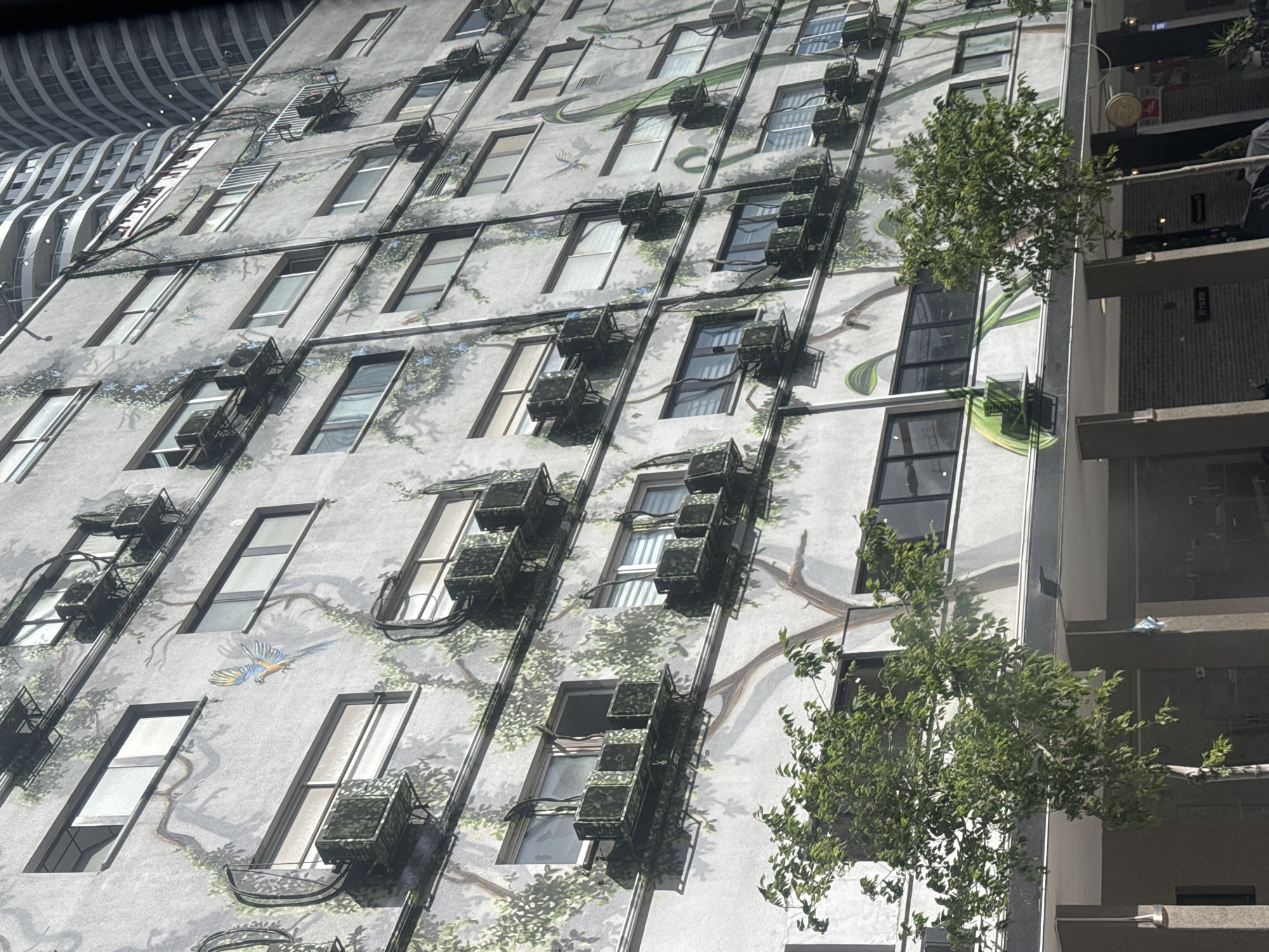The image shows the facade of a building in Tel-Aviv, Israel. The building features a mural depicting a whimsical scene of trees, leaves, and some animals (a snake and a bird) painted on its exterior. Many windows are visible, each with external air conditioning units attached. Adjacent to this building is another building with a more modern architectural style, including a balcony. The buildings are located in an urban area.