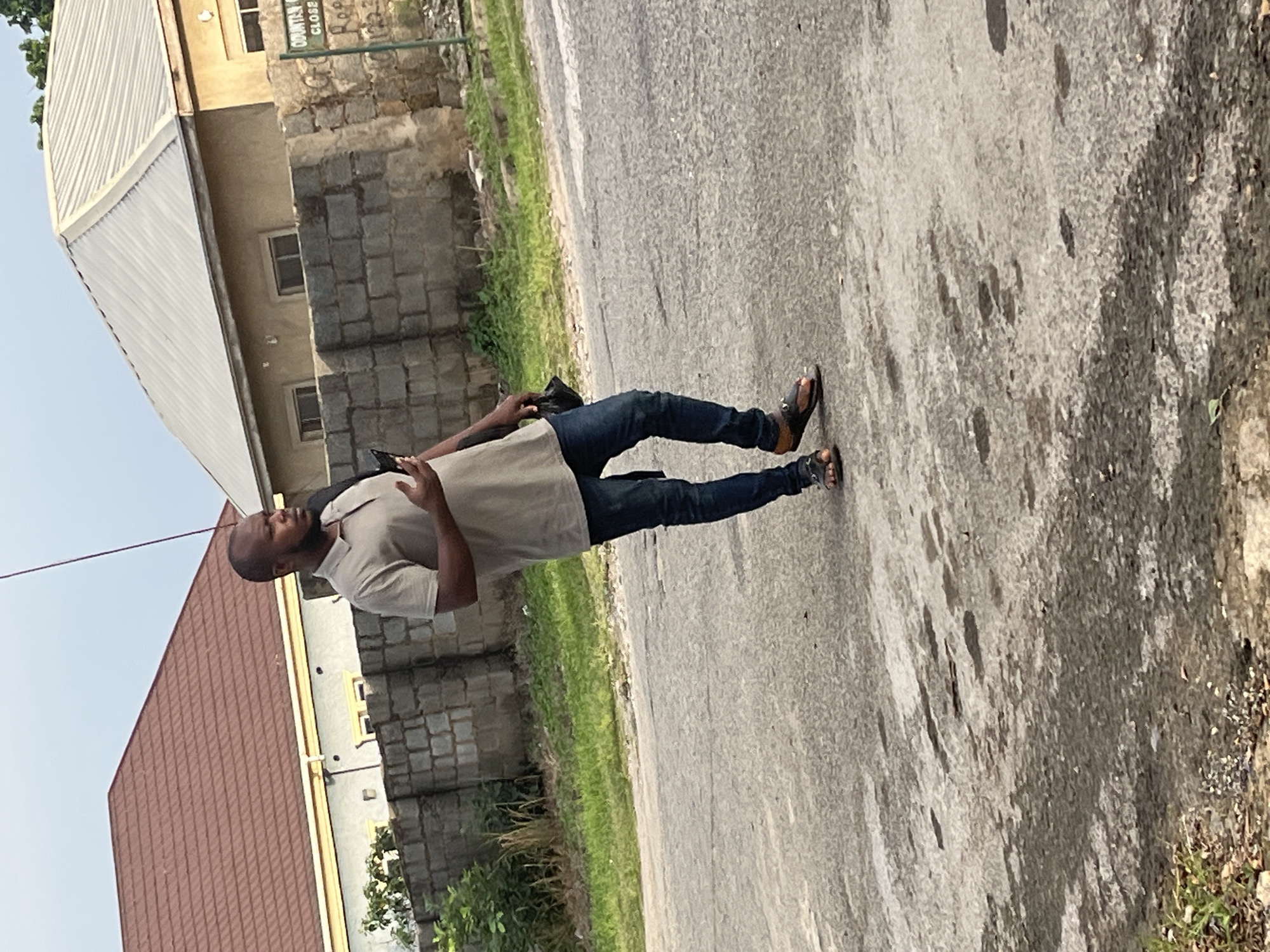 A man is standing on a paved road in Abuja, Nigeria, looking to his right while holding a phone in his right hand and a black pouch in his left. He is wearing a light brown polo shirt, blue jeans, and brown and black sandals. Behind him, a low brick wall separates the road from a grassy area and two buildings. The building on the left has a red tiled roof and light-colored walls with yellow trim. The building on the right has a silver corrugated metal roof and light brown walls, with a green sign that reads "ODUNTAN CLOSE". The sky is overcast.