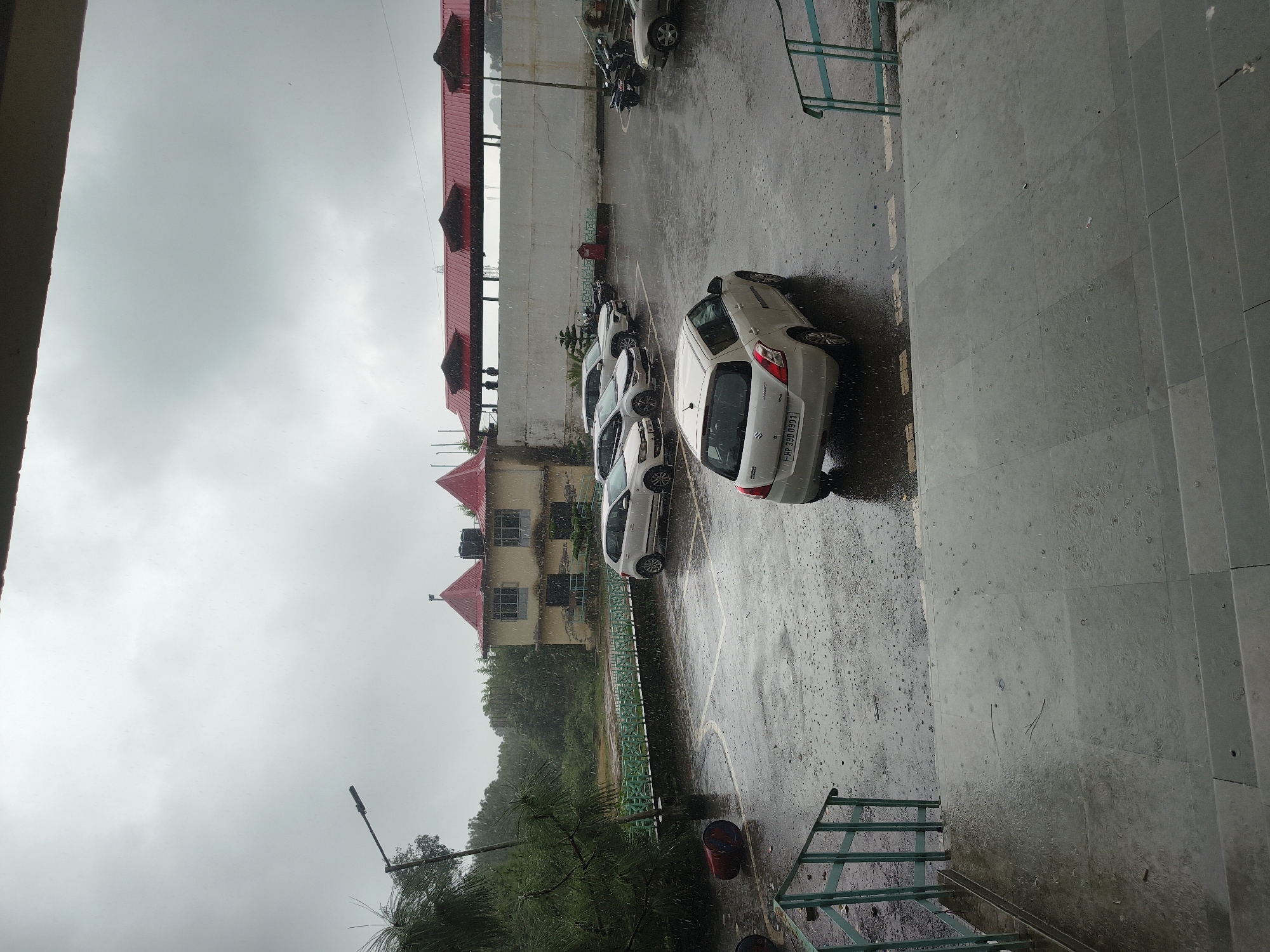 The image captures a scene in Dharamshala, India, during a rain shower. In the foreground, a grey tiled staircase leads down to a wet, paved area. To the left of the stairs, a green metal railing runs alongside. The paved area is a parking lot with several cars parked, including a white Maruti Suzuki Swift with license plate "HP 390 0901" in the foreground, facing away from the viewer. Further back, more cars are visible, including a white Volkswagen Polo and a white Toyota Fortuner. The ground is wet and reflective, with visible raindrops creating ripples. A green metal fence borders the parking lot, and beyond it, a building with a yellowish facade and red-tiled roofs stands. Trees and foliage are present in the background. The sky is overcast and grey, indicating the rainy weather. A street light is visible on the left side of the image. The scene appears to be an outdoor parking area adjacent to a building complex.
