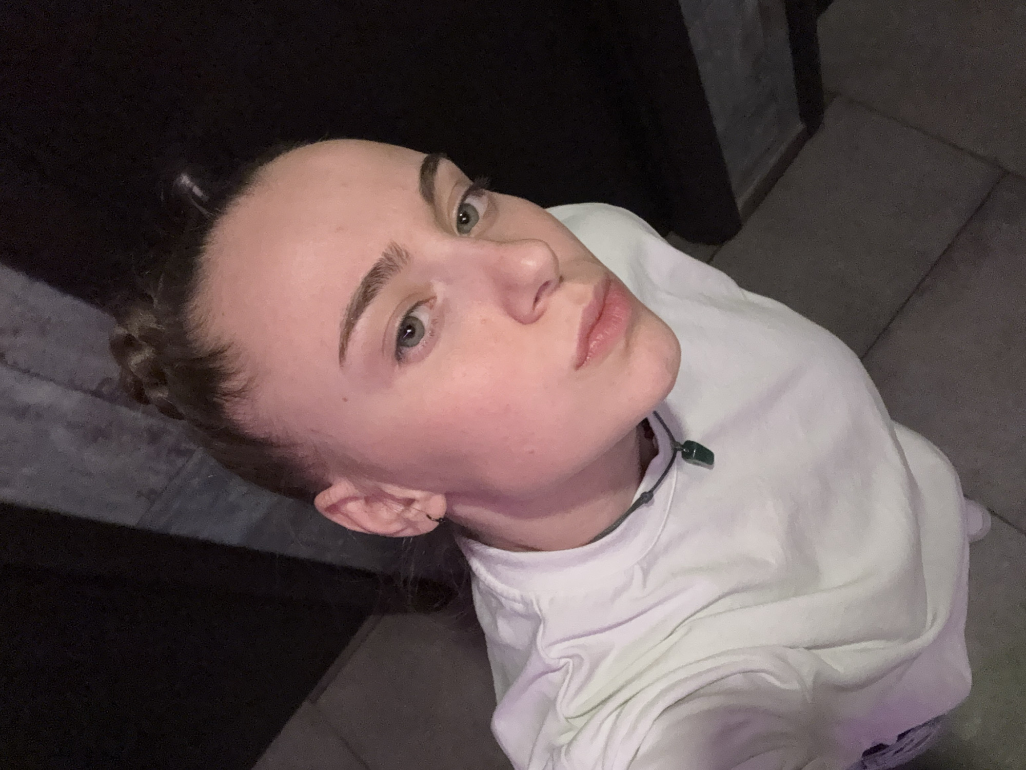 A person is photographed from a low angle, gazing upward toward the camera. They appear to be fair-skinned with light eyebrows and braided hair. The individual is wearing a light-colored t-shirt and a necklace with a dark stone pendant. They have an earring in their left ear. The background consists of dark walls and gray tiled flooring. The location of the image within Sofia, Bulgaria cannot be determined from the visible information in the image.