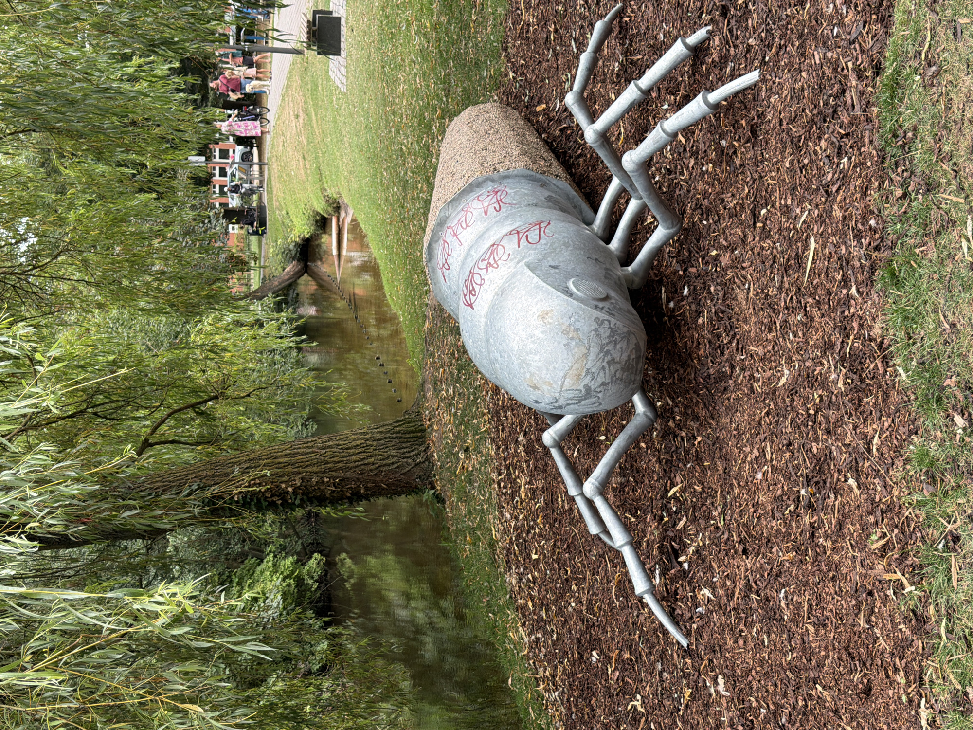 An outdoor sculpture of an insect lies on a ground covered in mulch, with grass in the foreground. The sculpture is made of metal and has a textured, light-brown segment at one end, and a shiny metal segment with red graffiti markings at the other. Six metallic legs extend from the body, positioned on the mulch. Behind the sculpture, a body of water, possibly a canal or river, is visible, bordered by a grassy bank. A weeping willow tree with long, flowing branches overhangs the water and extends into the upper left portion of the image. In the background, beyond the grassy bank and walkway, a park-like setting is evident with people walking, some with bicycles and dogs, and a few parked cars. The overall scene suggests a public outdoor space with a decorative art installation.