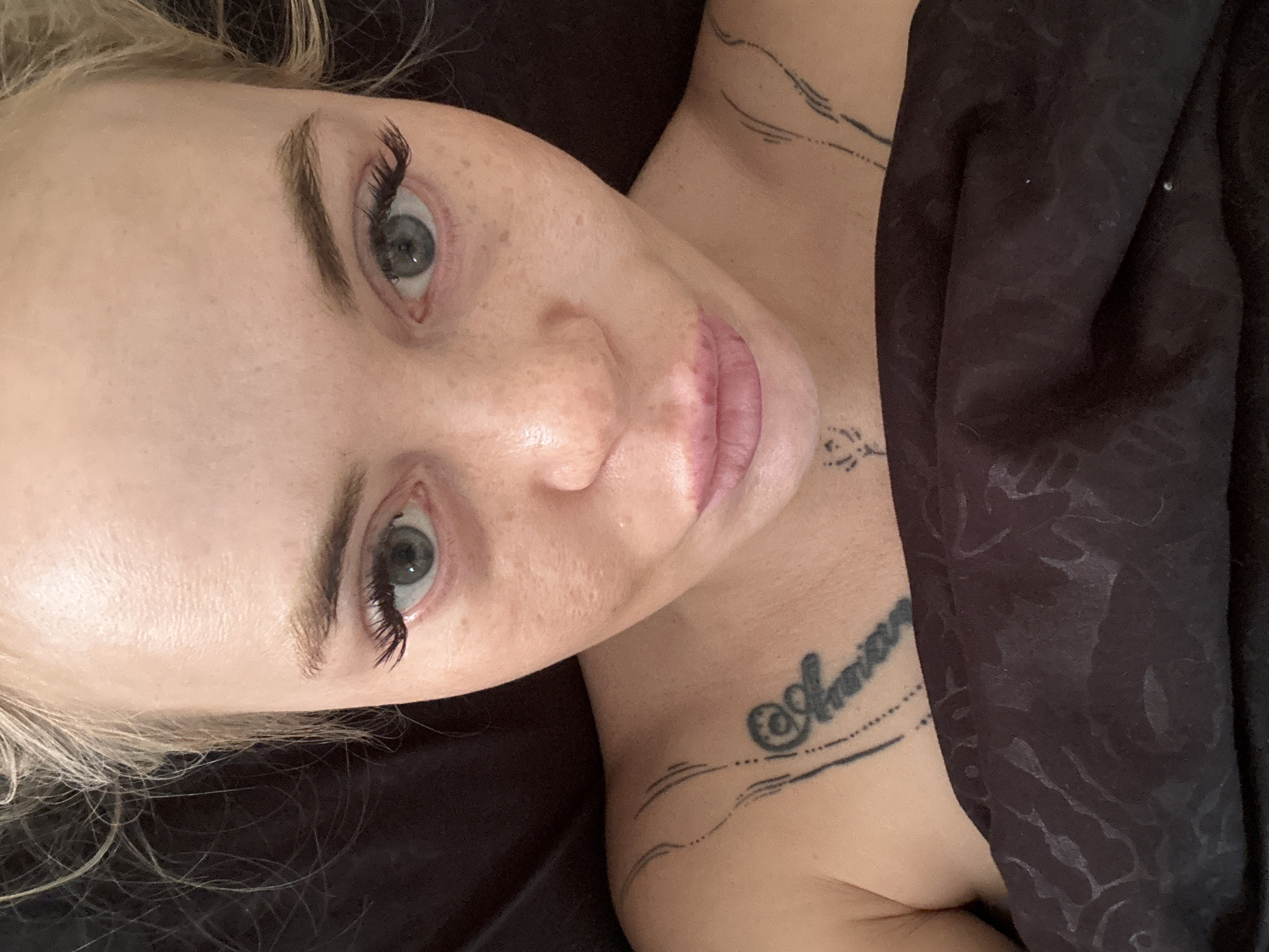 A close-up, overhead shot shows a pale-skinned woman with blonde hair and light green eyes. She has freckles across her nose and cheeks, and her eyes are slightly puffy. Her eyebrows are dark and defined, and her eyelashes are long and dark. She is wearing light pink lipstick, and her lips are slightly pursed. There are several black tattoos on her upper chest and shoulder. She is wearing a dark-colored, textured fabric that covers her chest. The background is dark and out of focus.
