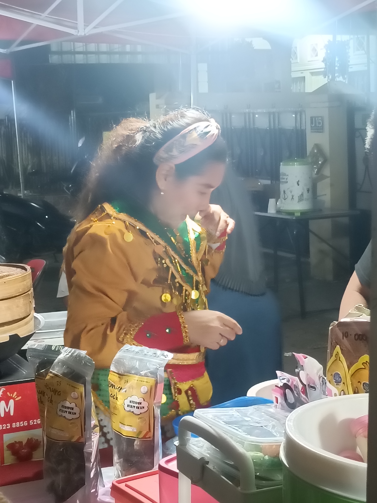 A woman in traditional Indonesian attire, adorned with gold sequins and a green sash, stands at a market stall in the city of Kediri, Indonesia. She is seen looking down at a small object she is holding, possibly a food item or a coin, with her right hand. The stall displays various packaged goods, including what appears to be fish skin crackers, with visible labels. In the background, a white tent structure with lighting is present, suggesting an outdoor market setting. There are also other individuals and market stalls visible in the blurred background, indicating a lively commercial environment. The overall scene captures a moment of transaction or preparation at a local marketplace.