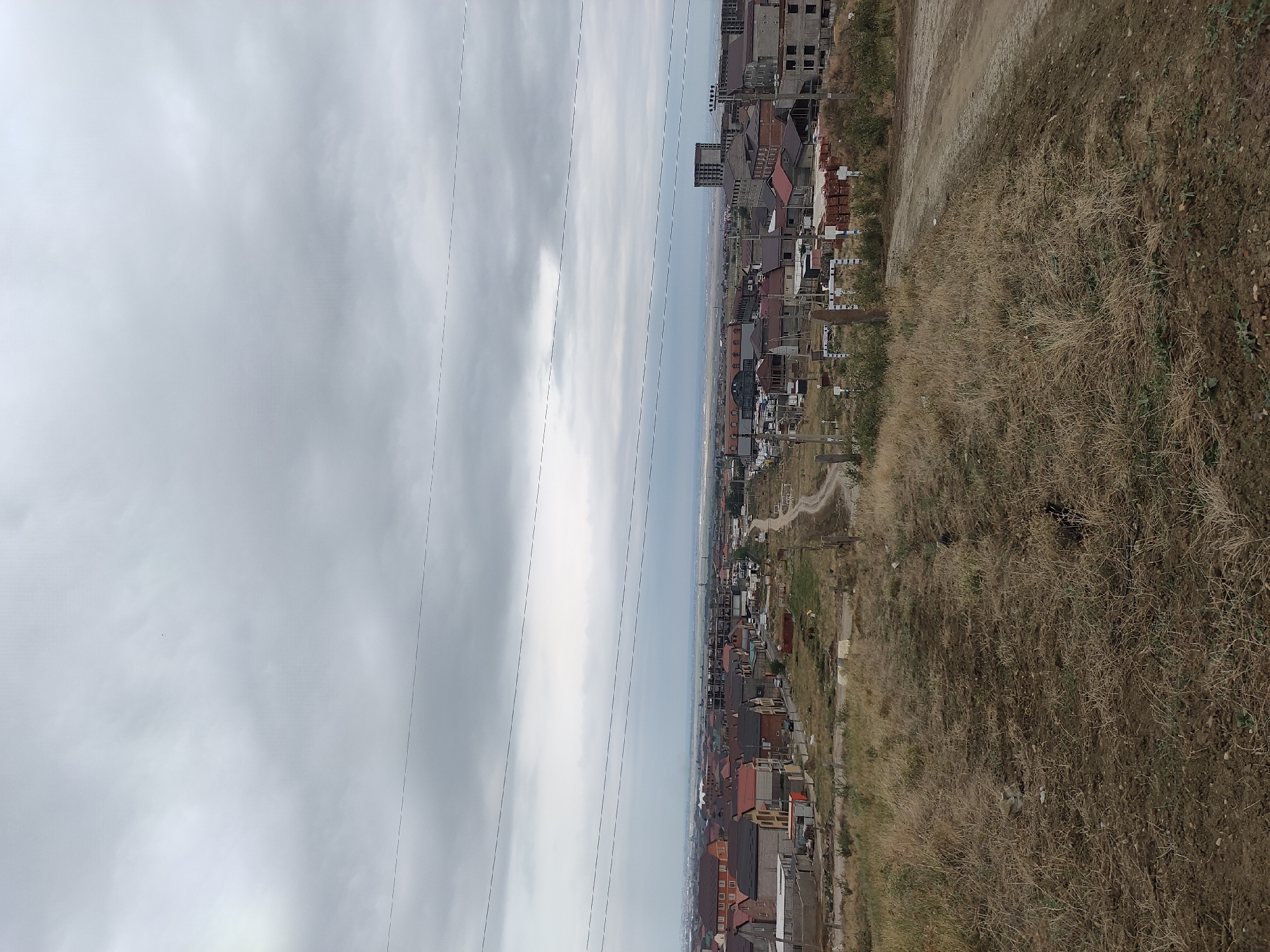 An exterior, high-angle, eye-level, full shot captures a wide view of a residential area on a cloudy day. The sky is overcast, with a faint haze obscuring the distant horizon. Power lines crisscross the upper portion of the frame. In the foreground, dry, brown grass covers the uneven terrain, with a dirt path winding its way through it. Below, a developing neighborhood unfolds, featuring a mix of completed homes with red and brown roofs, and buildings under construction with visible concrete structures. There are stacks of bricks, construction materials, and some vehicles scattered throughout the area. A few small animals are visible in the fields among the houses. The scene suggests a rural or semi-urban environment undergoing development.