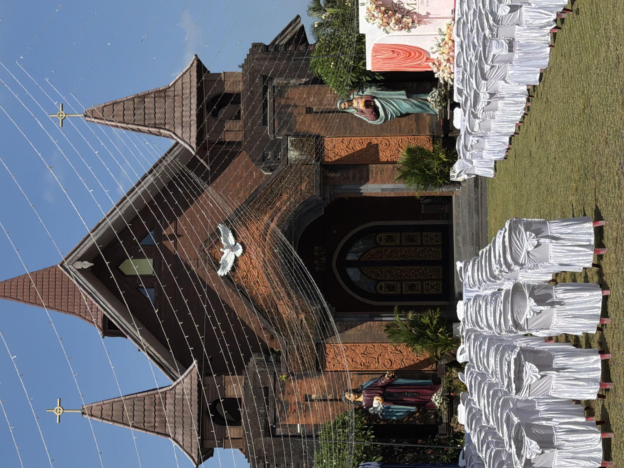 This is a ground-level, eye-level, medium shot of the facade of a Roman Catholic church in North Kuta, Indonesia, on a clear day. The church is constructed of reddish-brown brick, with two prominent pointed towers topped by gold crosses. A series of white string lights are draped across the front of the building and between the towers, some appearing to have small bulbs or decorations. A white dove sculpture is positioned above the central entrance. Flanking the entrance are two life-sized statues: on the left, a male figure in a purple and blue robe holding a white dove, and on the right, a female figure in a green robe holding her hands to her chest. The main entrance is a large, arched doorway with decorative golden doors.
In front of the church, rows of white-covered chairs are neatly arranged on a green lawn, suggesting an outdoor event or ceremony. To the right of the entrance, a white banner with floral decorations and text, partially obscured, is visible, further indicating a planned gathering. The scene is devoid of people. The environment appears to be well-maintained, with lush green grass and some surrounding foliage.