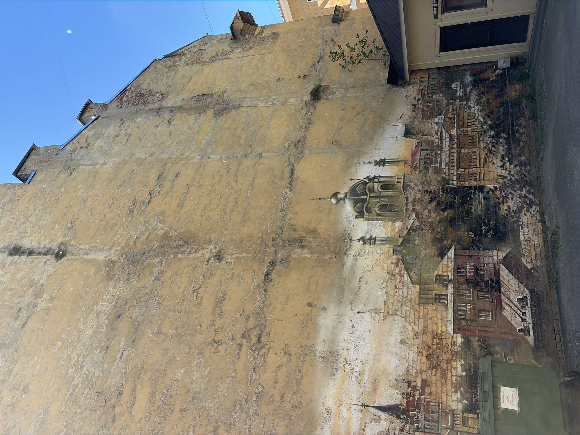 The image depicts a mural on the exterior wall of a building, likely in Saint Petersburg, Russia, given the architectural style of the painted city. The mural features a panoramic view of a city with numerous buildings, spires, and domes, rendered in a somewhat aged and weathered style. The dominant colors in the mural are muted yellows, browns, and greys, suggesting a historical or old-world setting. The actual wall itself is textured and shows signs of wear, with peeling paint and visible brickwork, which adds to the aged aesthetic of the artwork. In the foreground, to the left, a large green dumpster is visible, with a small white notice attached to it. To the right, a portion of a small, modern structure with a dark door and window can be seen, contrasting with the mural. The sky above is a clear, bright blue. The overall impression is of a public artwork integrated into the urban landscape, depicting a historical cityscape on a weathered building.