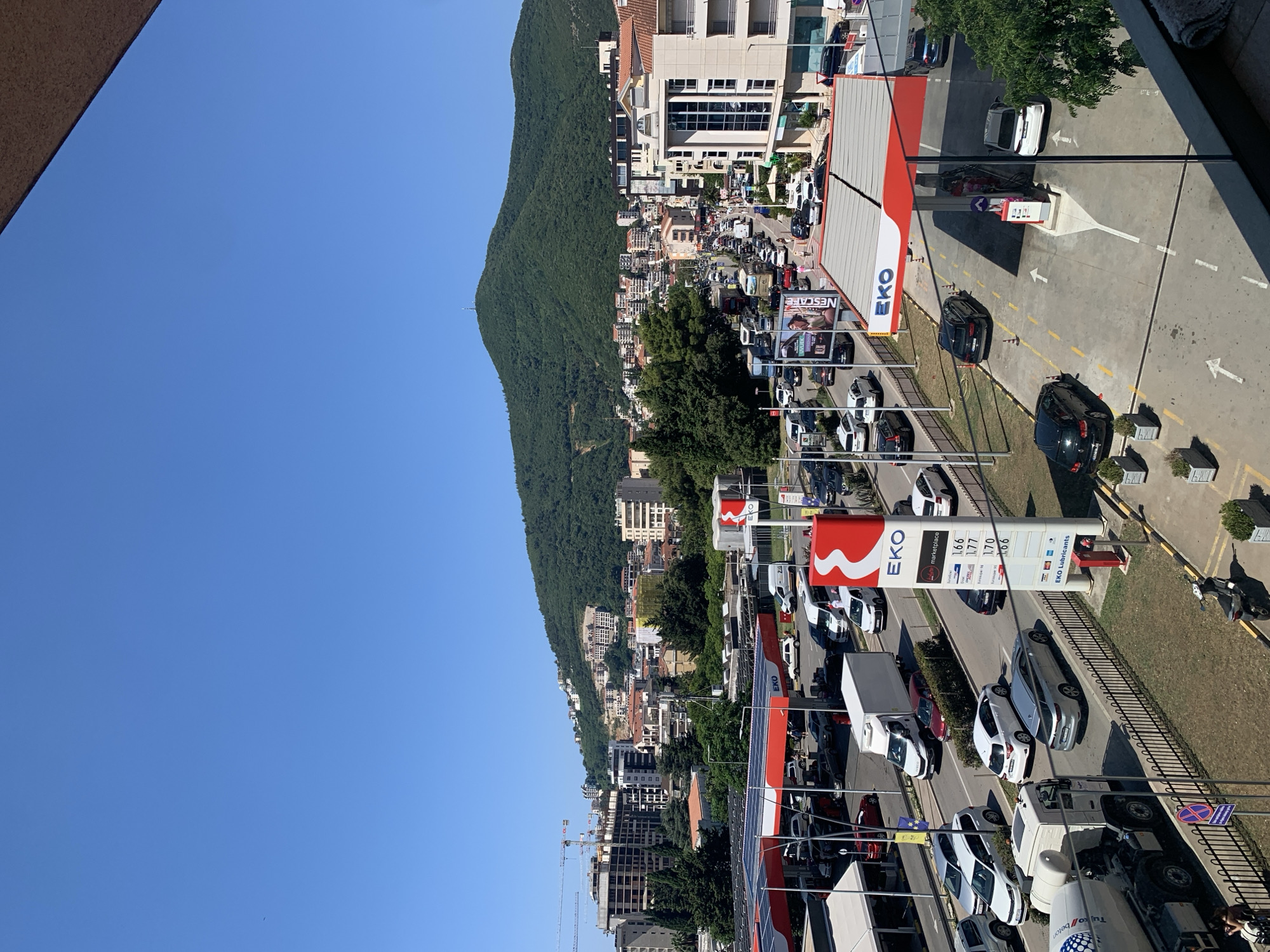 The image shows a view of a busy street and a gas station in Budva, Montenegro, with a lush green hill in the background. The sky is clear and blue.
In the foreground, a gas station with red and white canopies is visible, with several cars filling up their tanks. A prominent "EKO" sign stands tall, displaying fuel prices. Adjacent to the gas station, a busy road is lined with vehicles, including cars, vans, and trucks. Some construction is ongoing in the middle ground, with cranes visible near unfinished buildings.
The middle ground of the image features a dense urban landscape with apartment buildings and shops. Trees line the streets, providing shade. The background is dominated by a large, verdant hill dotted with buildings, some appearing to be residential. A telecommunications tower is visible on the crest of the hill.
People are seen at the gas station and walking along the sidewalks. The overall scene depicts a vibrant, active urban environment under a bright, sunny sky.