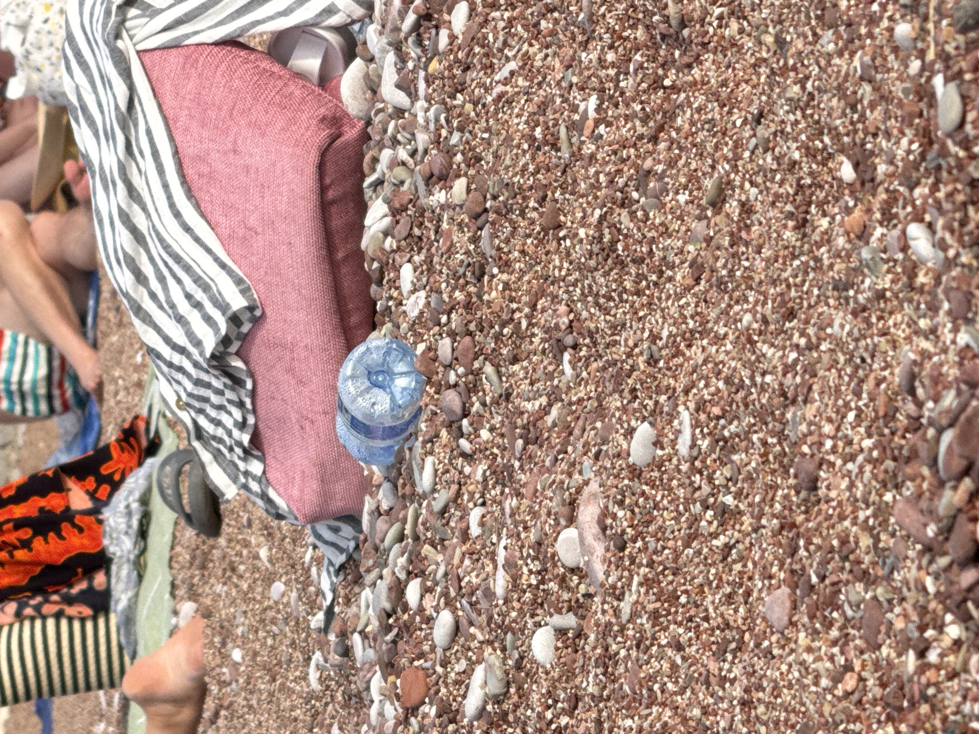 The image depicts a detailed close-up of a pebbly beach in Sveti Stefan, Montenegro, with blurred figures in the background. The foreground is dominated by a dense surface of small, reddish-brown and lighter-colored pebbles.
In the mid-ground, resting on the pebbles, is a large, rectangular, textured pink or mauve textile item, possibly a beach bag or cushion. Draped over this item is a white and dark gray horizontally striped textile, likely a towel or shirt. To the right of the pink item, a single light-colored sandal is partially visible. To its left, a dark-colored sandal is also present on the pebbles. Two partially filled clear plastic water bottles lie horizontally on the pebbles to the left of the main textile items.
The background features several out-of-focus individuals relaxing on the beach. Visible elements include a bare foot resting on a light green mat, striped fabric (yellow/beige and dark stripes), and an orange and black patterned fabric, suggesting beach attire or towels. The people appear to be seated or lying down. The scene is illuminated by natural daylight.