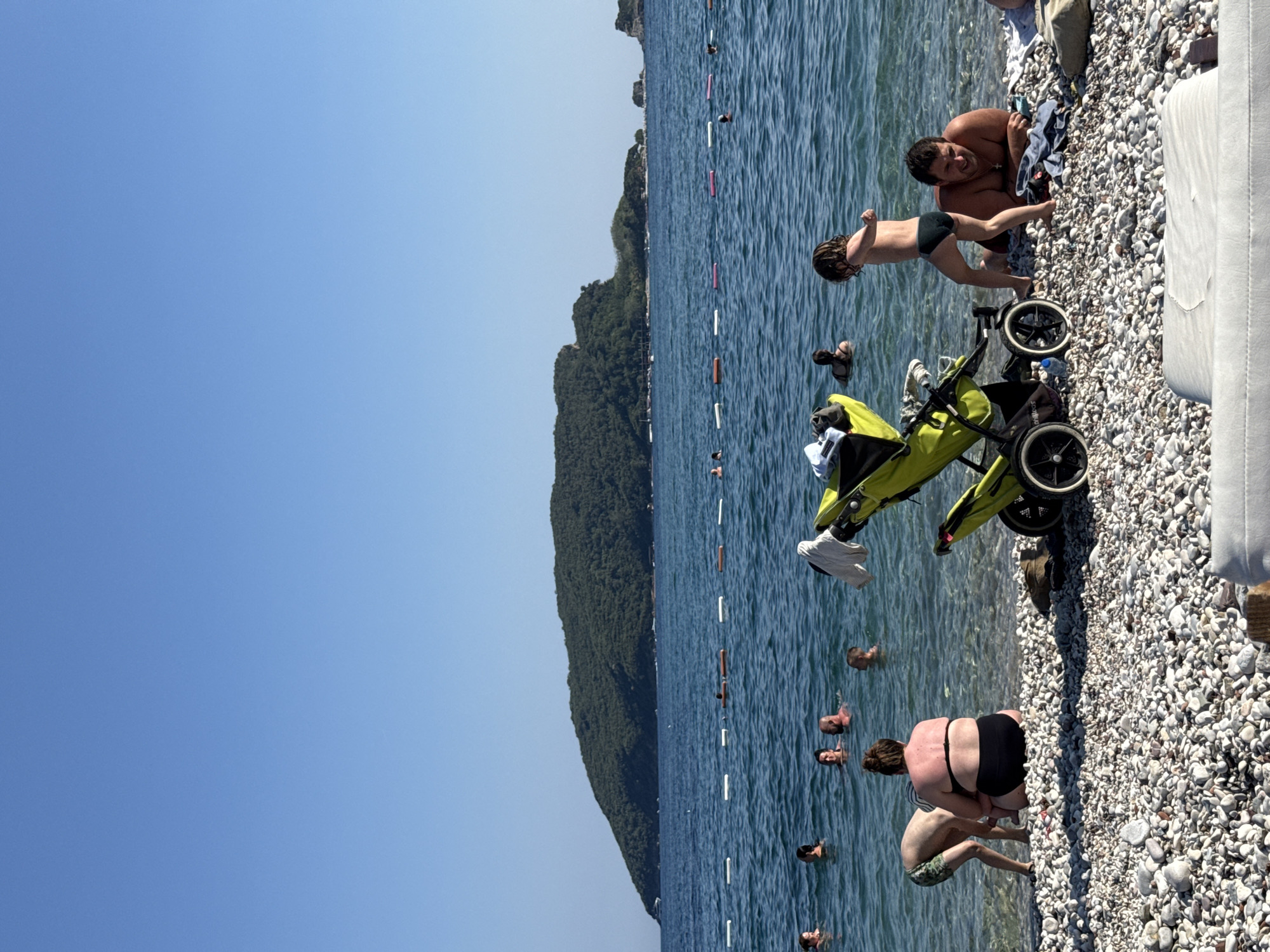 This is a photograph taken from a beach in Budva, Montenegro. The scene depicts a sunny day at the beach, with a clear blue sky and calm, clear water. Several people are visible in the water, some swimming and others wading. On the pebbly shore, a man is lying down and looking at the camera, with a young child in swimwear standing next to him. A neon green stroller is parked nearby on the pebbles. In the background, a lush, tree-covered mountain rises from the sea, and a row of buoys marks a swimming area. The overall atmosphere is relaxed and summery. The time of day appears to be midday, given the bright sunlight.