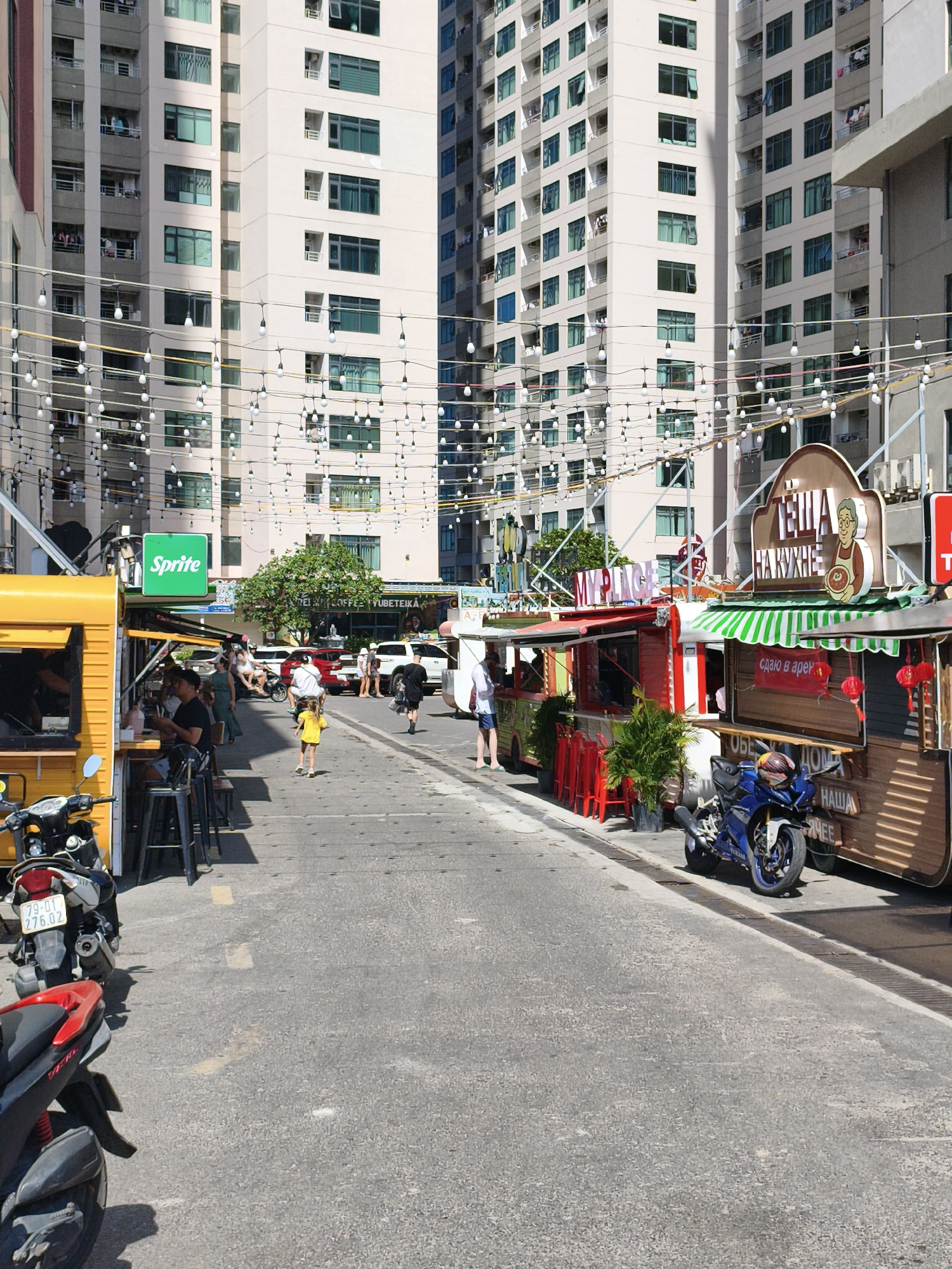This is a street scene in Nha Trang, Vietnam, likely in a modern, urban area with tall residential buildings in the background. The street is lined with food stalls and small shops, decorated with strings of lights. Several people are visible, some walking, some sitting at outdoor tables, and others working at the stalls. Two motorcycles are parked on the side of the street. The overall impression is one of a lively street market or dining area.