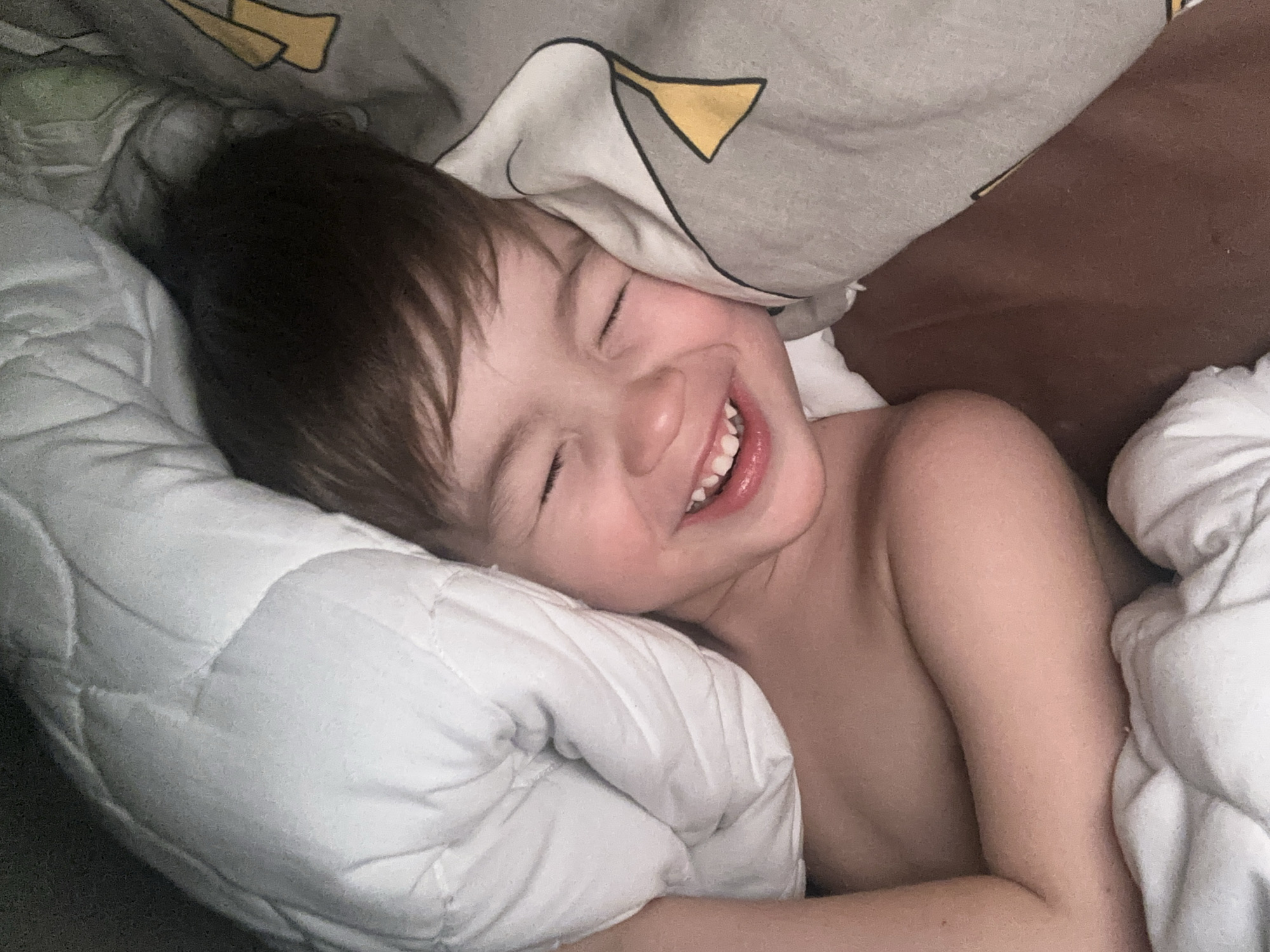 A young child is lying in bed, appearing to be sleeping or resting with a smile on their face. The child is nestled between a white quilted blanket and a grey patterned blanket with a yellow design. The child's eyes are closed. The location, Khimki, Russia, is not visually discernible from the image.