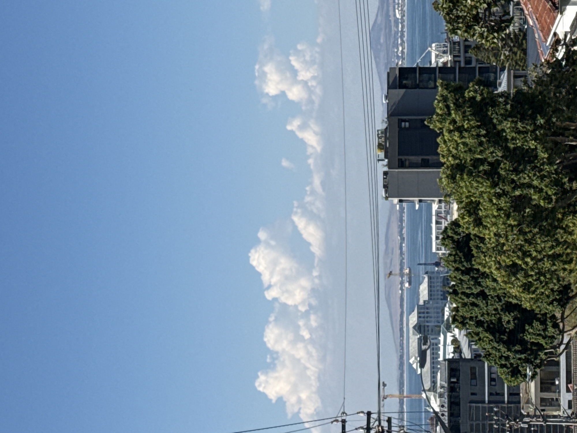 The image is a view from a hillside in Cape Town, South Africa. The sky is blue with cumulus clouds. Power lines cross the view. A body of water, likely the Atlantic Ocean, is visible with distant hills on the opposite shore. Buildings and trees are on the right side of the image.