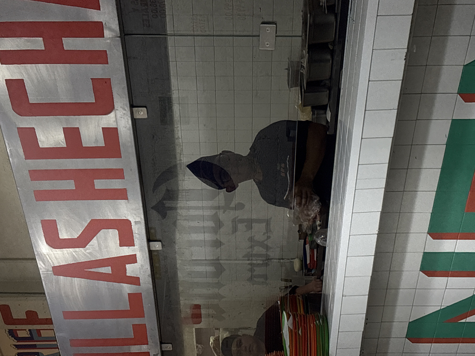 The image shows a reflection of a person wearing a blue hairnet and a gray t-shirt inside a business. The business name "TALLAS CHECK" is prominently displayed in large red letters on a silver surface. The reflection suggests that the person is working behind a counter or preparation area. There are items such as plastic bags and containers visible on the counter. The background consists of a tiled wall and a partial view of another sign with green and red stripes. The reflection of arrows and the word "EXIT" on the glass provide a clue to the location. Based on the business name and the visual cues, it is likely that the image was taken in a business located in Mexico City, Mexico.