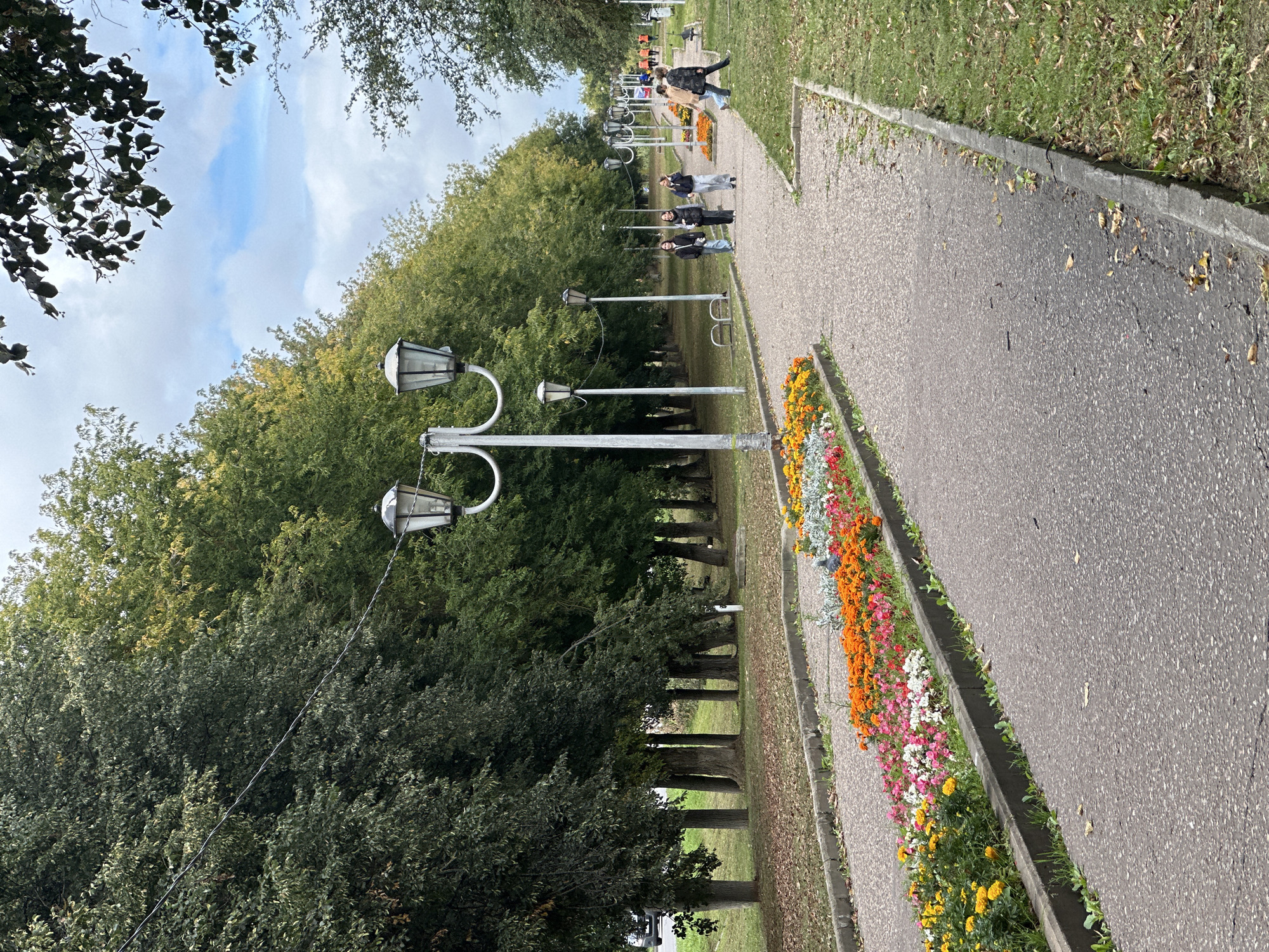 A wide paved walkway, flanked by flowerbeds on the left and a grassy area on the right, recedes into the distance. Several vintage street lamps are spaced along the walkway. A line of mature trees with dense foliage runs along the far side of the walkway, casting shadows. The sky is overcast with patches of blue visible. Several people are walking along the path. In the foreground, a flowerbed bursting with vibrant orange, pink, and yellow flowers runs parallel to the walkway. The scene appears to be in a park or public garden in Chekhov, Russia.
