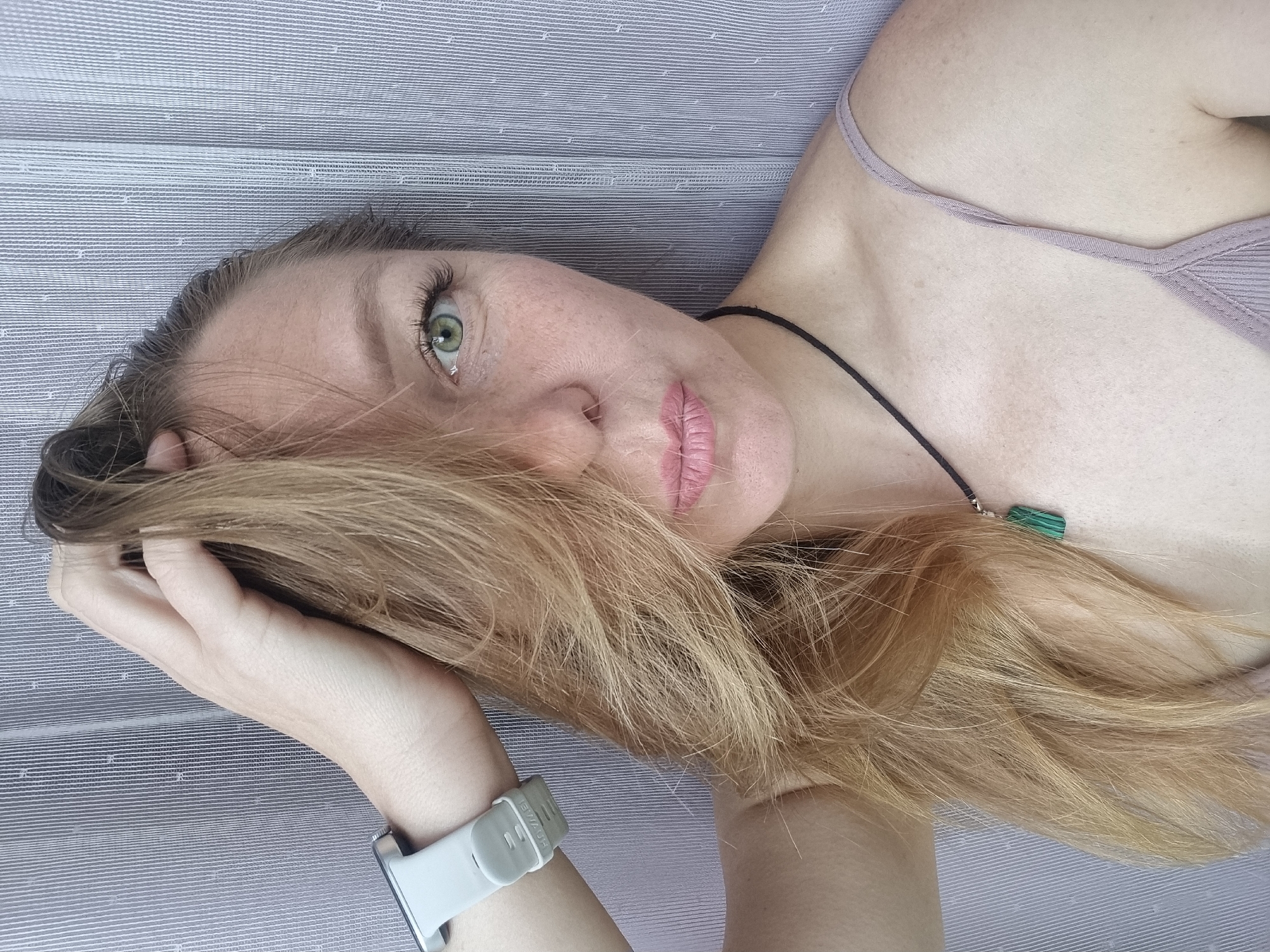 A selfie of a fair-skinned woman with blonde hair and green eyes is taken against a striped, grey-blue backdrop. She is wearing a light-colored tank top and a black cord necklace with a green pendant. A smartwatch with a light grey band is visible on her left wrist. Her hair is pulled back with her hand, obscuring part of her face. The image appears to have been taken indoors, possibly in a residential setting. There is no discernible location information within the image that would indicate it is from Chaikovsky, Russia.