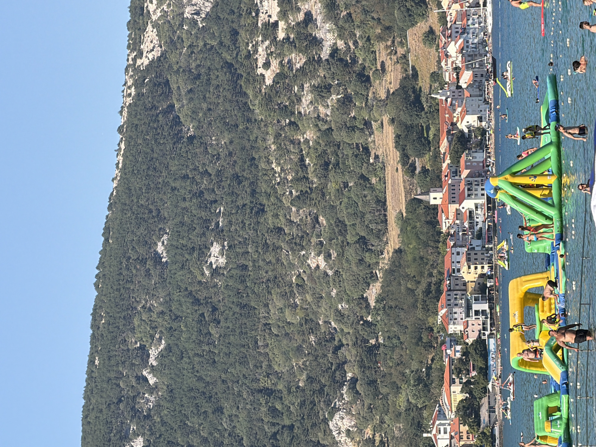 The image depicts a sunny day at the beach in Općina Baška, Croatia. In the foreground, a vibrant inflatable water park dominates the scene, with various colorful structures like slides and climbing walls. Several people are actively engaged on and around the water park, some climbing, others playing, and some wading in the shallow water. In the mid-ground, a charming coastal town with red-tiled roofs and white buildings lines the waterfront. Boats are visible anchored in the bay, and other watercraft, including what appear to be pedal boats, are present. The background features a steep, densely forested hill that rises majestically behind the town, with patches of rocky terrain visible amidst the trees. The sky is a clear, bright blue, indicating fair weather.