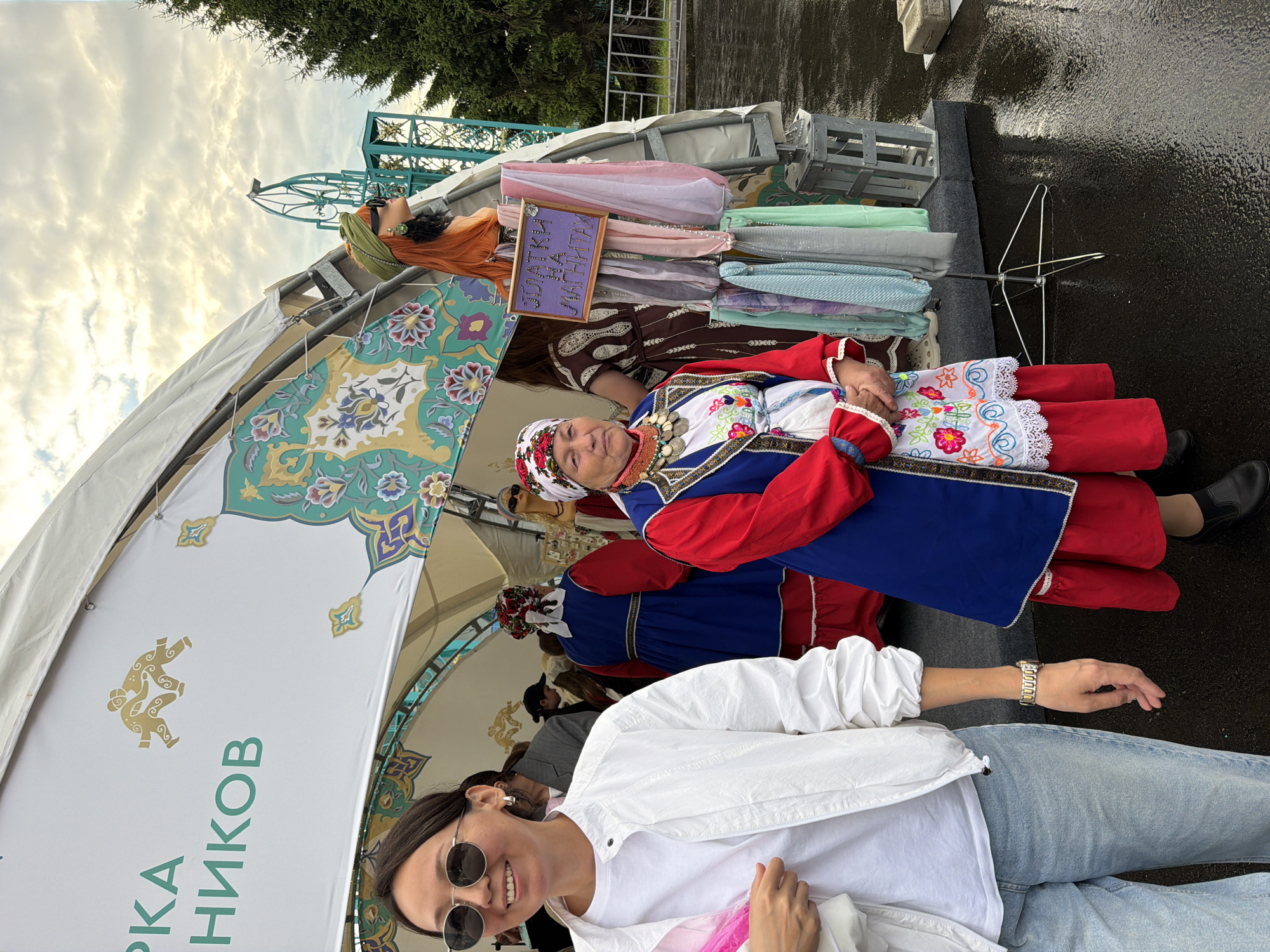 An outdoor scene features a woman in the foreground wearing a white jacket, white t-shirt, and light blue jeans, accessorized with sunglasses and a watch. To her right, an older woman in traditional ethnic attire, including a red and blue dress with intricate embroidery and a red headscarf, stands prominently. Behind them, another woman in a similar blue and red traditional dress is visible. A stall displaying scarves and accessories is also present, with a mannequin head wearing a green headscarf. The background includes a tent with decorative patterns and text, a tree, and what appears to be part of an amusement park ride. The ground is wet, suggesting recent rain. The overall atmosphere is that of a cultural fair or market.
