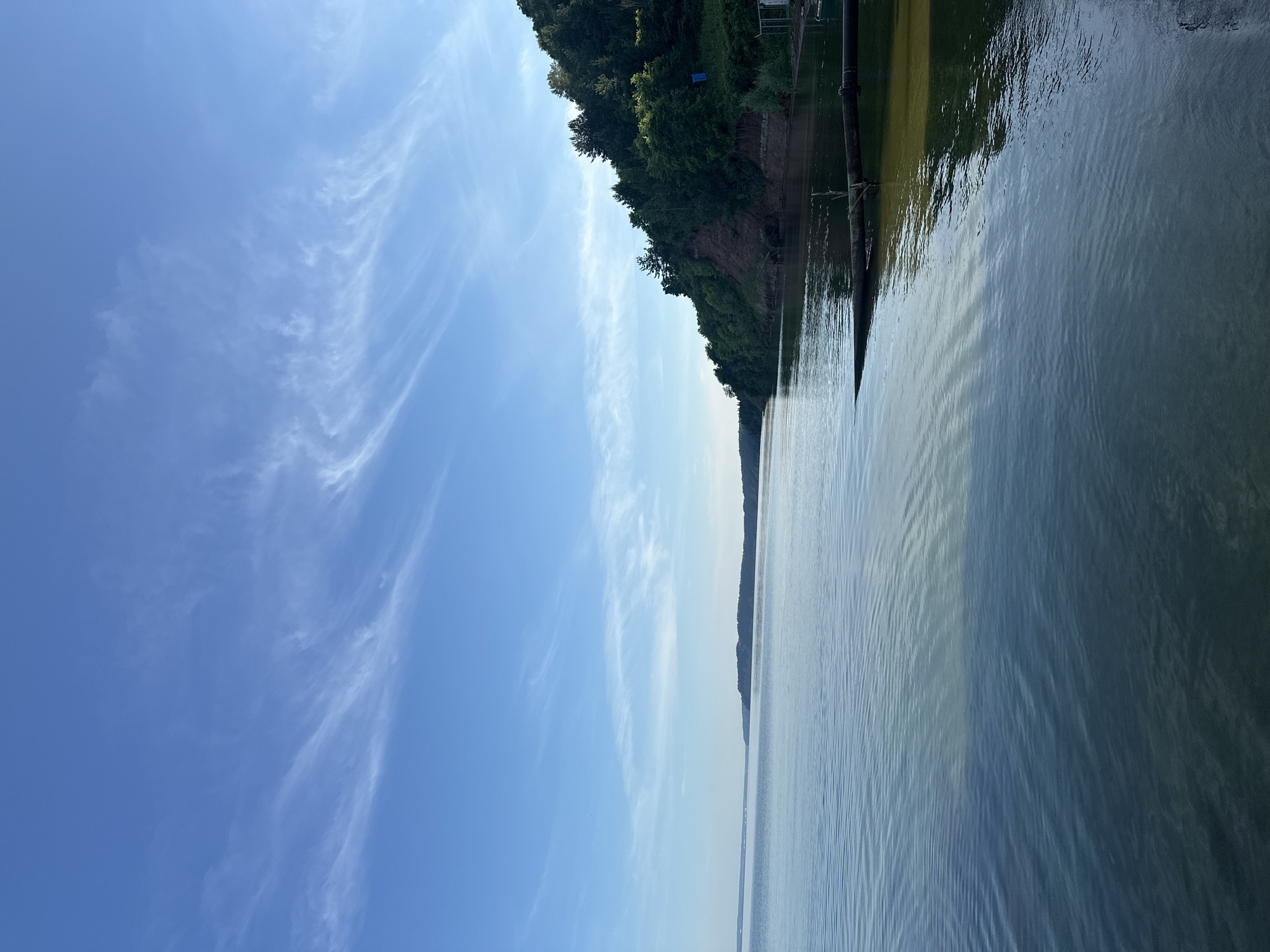 The image depicts a serene view of a wide body of water, likely a river or lake, with a lush green, tree-covered embankment on the right side. The sky is a clear blue with wispy white clouds stretching across it. The water's surface is calm, reflecting the sky and surrounding landscape, with gentle ripples visible. Along the embankment, there are areas of exposed earth and dense foliage, suggesting a natural, undeveloped area. In the foreground, a large, dark pipe or log extends into the water from the right bank. On the embankment, a small blue structure and parts of what appears to be a fence or scaffolding can be seen. The overall atmosphere is peaceful and natural, characteristic of a scenic riverside location. This image is likely taken near the city of Galovo, Russia, given the geographical context.