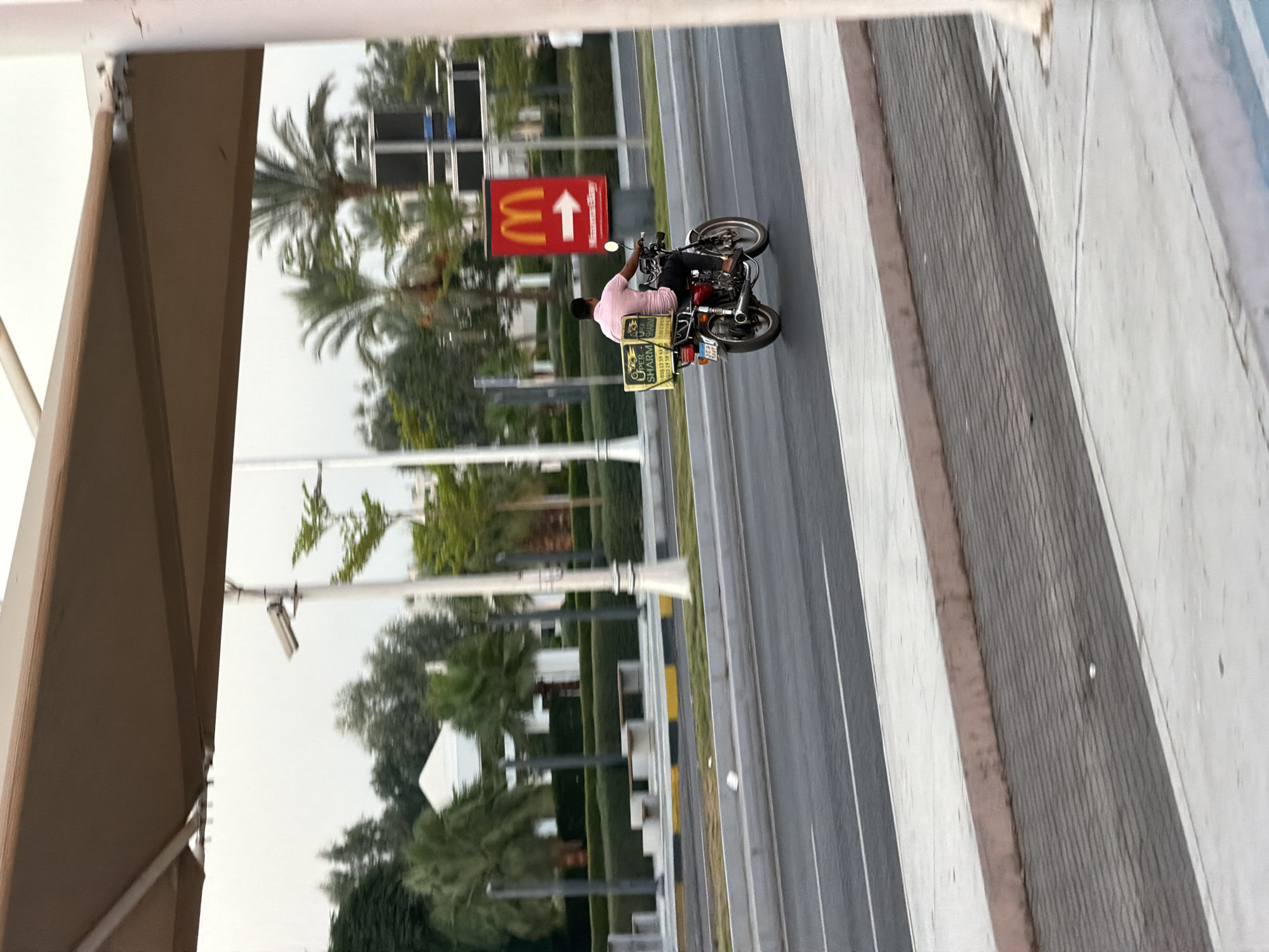 A man wearing a pink shirt and black pants is riding a red motorcycle on a multi-lane road in Sharm El Sheikh, Egypt. The motorcycle has a yellow delivery box on the back with text that is partially visible. In the background, there is a McDonald's sign with an arrow pointing to the right. There are also palm trees, bushes, and buildings, suggesting an urban or suburban environment. The sky is overcast, indicating a cloudy day. The overall scene appears to be a typical street view in a warm climate.