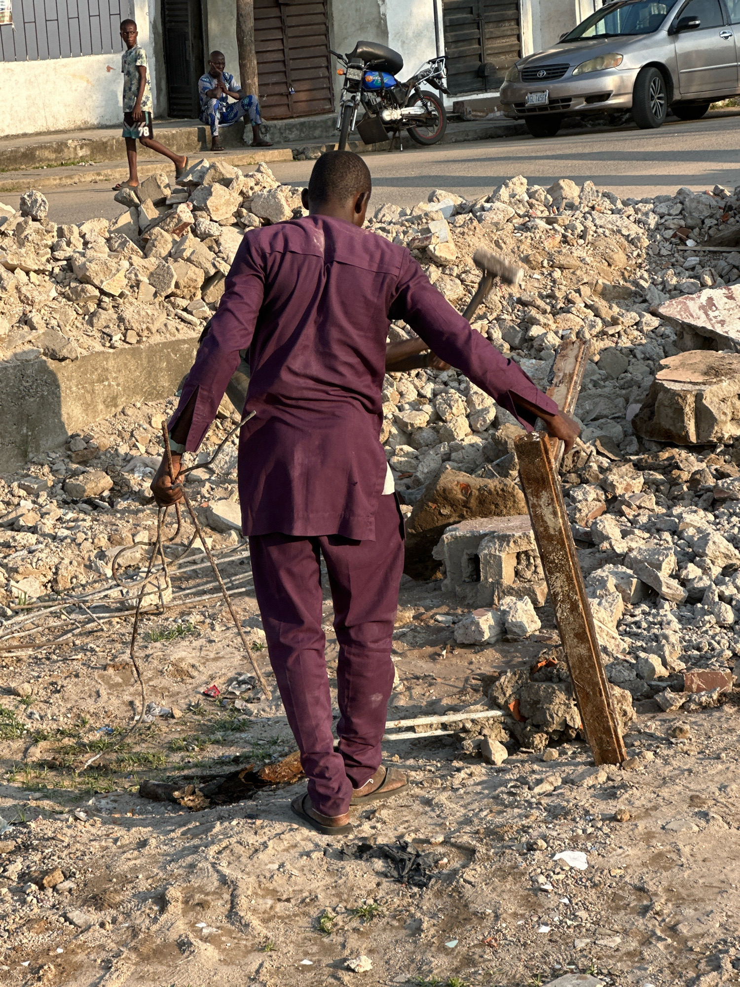 The image depicts a street scene in Badagry, Nigeria, characterized by debris and construction materials. In the foreground, a man in a purple traditional outfit is holding a bundle of bent metal rods in his left hand and a long, rusted metal beam in his right hand, which he appears to be clearing. He is facing away from the camera, towards a pile of rubble composed of broken concrete, bricks, and stones. Behind him, a motorcycle is parked, and a silver sedan is visible on the street. Further in the background, two young boys are present. One boy, wearing a patterned blue outfit, sits on a step, while another, in a t-shirt and shorts, walks in front of him. The environment suggests ongoing construction or demolition work, with the debris dominating the visual space.
