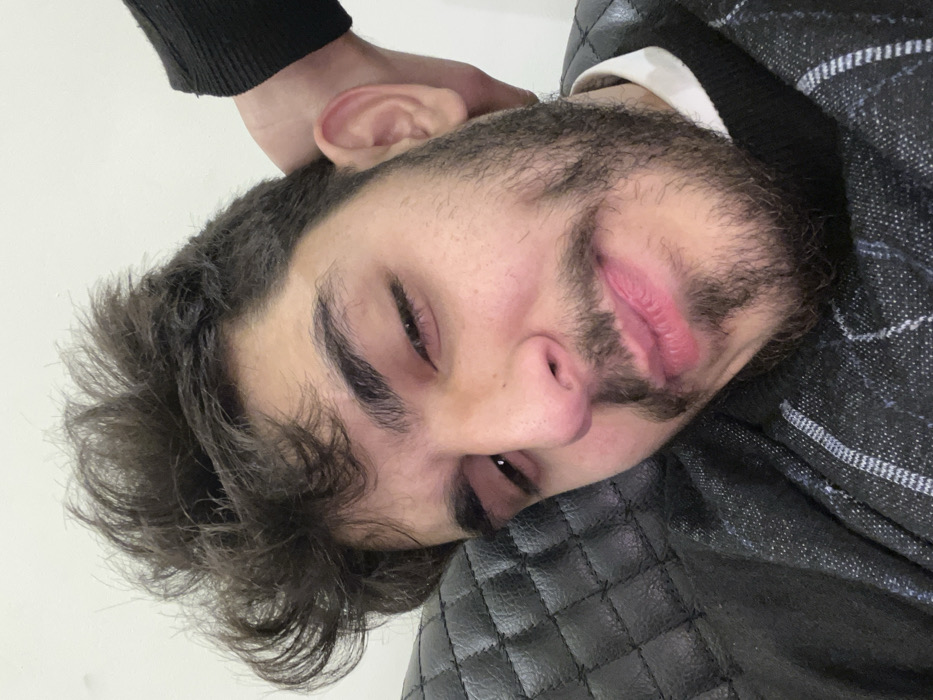 A close-up image depicts a young adult male in an indoor environment within the city of Rukn ad-Din Municipality, Syria. The subject has dark, unkempt curly hair, dark eyebrows, a mustache, and a full beard. His eyes are partially closed, and his lips are closed and slightly upturned at the corners. He is wearing a dark-colored, possibly knit sweater with a subtle geometric pattern, over a white collared shirt. The man's head is resting against a dark, quilted, cushioned surface, consistent with an upholstered chair or sofa. A plain, light-colored wall forms the background in the upper right. A hand, partially obscured by a dark sleeve, is positioned behind the subject's right ear.