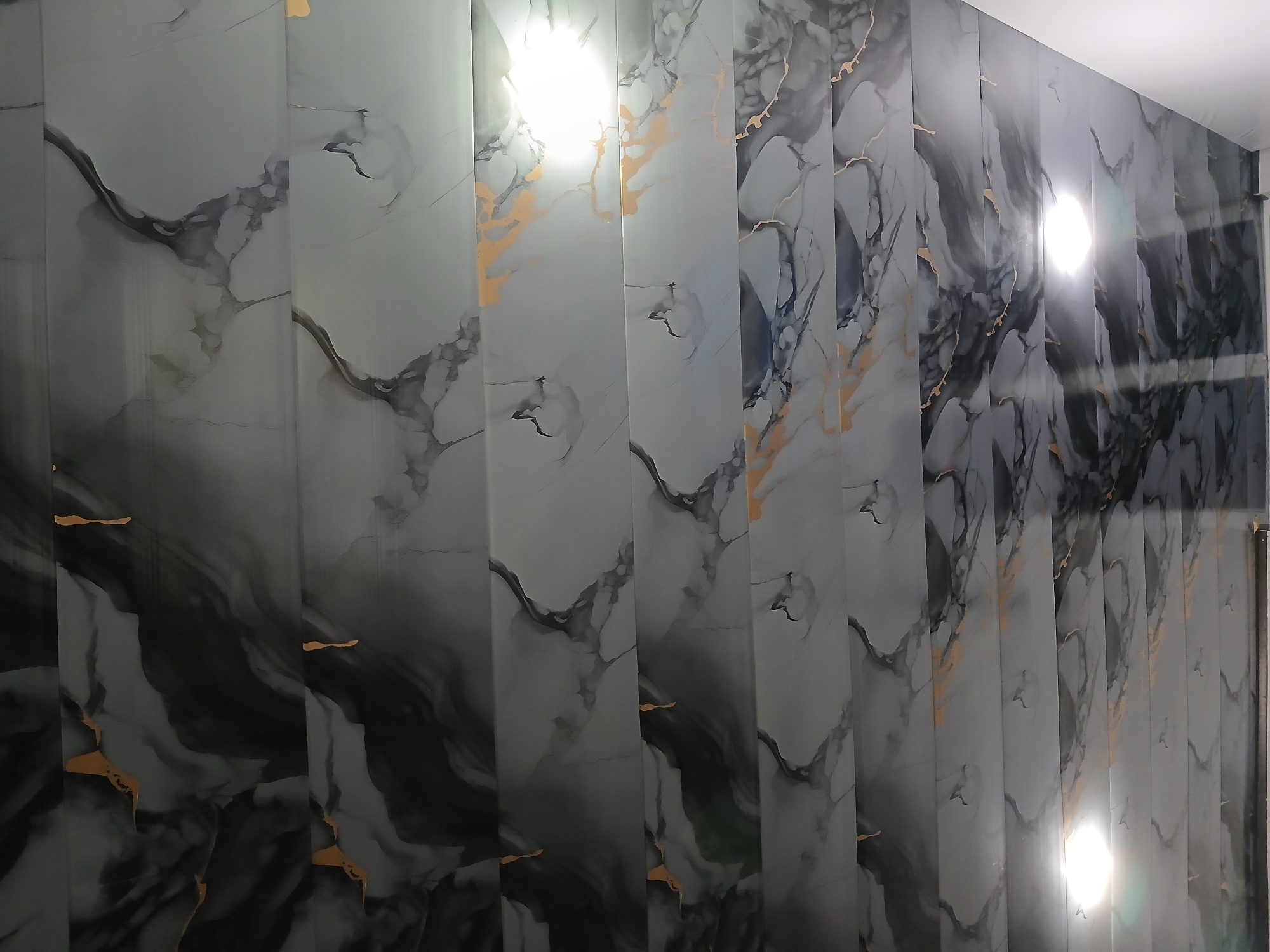 The image shows a ceiling with a marble-like pattern, featuring streaks of black, gray, and gold. The pattern is horizontal and divided into sections by visible lines, suggesting it is made of panels. There are two bright lights on the ceiling, one in the upper right section and another lower down. The overall impression is of an indoor space, possibly a room or hallway. The image does not contain any visual cues that would allow for the identification of its specific location within Xidirsho, Uzbekistan.