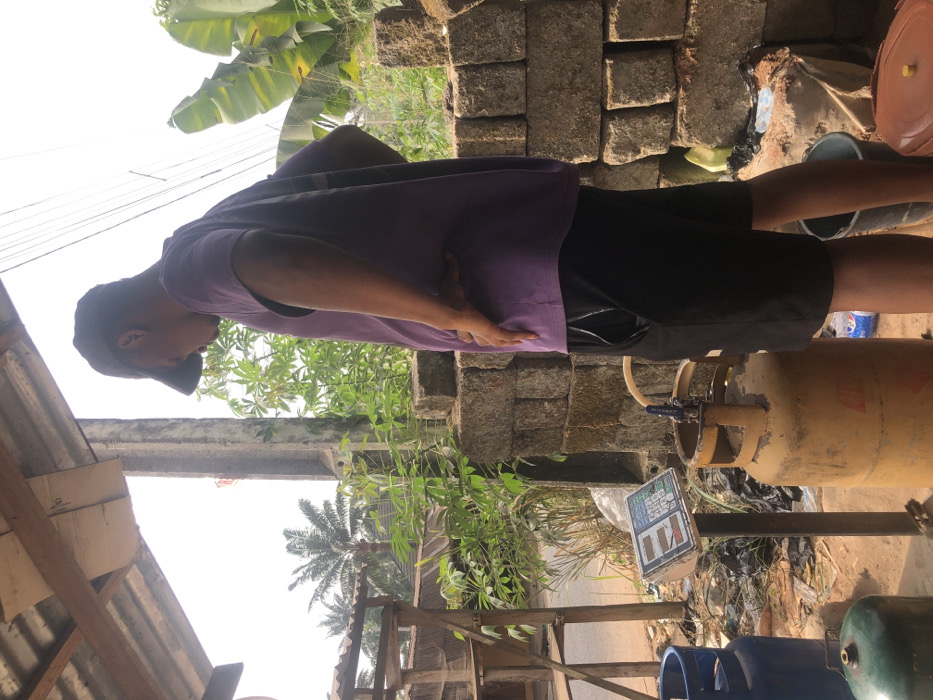 This image depicts an outdoor commercial scene in Alor, Nigeria, specifically a gas refilling or vending point.
A male individual, seen from the side facing right, stands centrally. He wears a purple short-sleeved shirt, black shorts, and a dark baseball cap, with his right hand on his hip and his head tilted downwards, observing the objects in front of him. To his left, a white digital weighing scale on a dark stand displays unreadable numbers. Directly in front of him and to the left of the scale is an upright, yellow liquid petroleum gas (LPG) cylinder with a valve and hose assembly attached to its top. Partially visible to the far left are a blue LPG cylinder and a green LPG cylinder on the ground.
The immediate ground is dirt, interspersed with some refuse, including plastic bottles and dark bags. In the background to the left, a wooden stall-like structure with shelving and a corrugated metal roof is visible, with a dirt road beyond it. Further back, dense green foliage and several palm trees are present. To the right of the individual, a stack of concrete blocks forms a wall, behind which more green vegetation is visible, including large-leafed plants. Overhead, a corrugated metal roof supported by wooden beams is partially visible, along with a concrete pillar. The scene is illuminated by bright daylight, under a clear or brightly overcast sky.