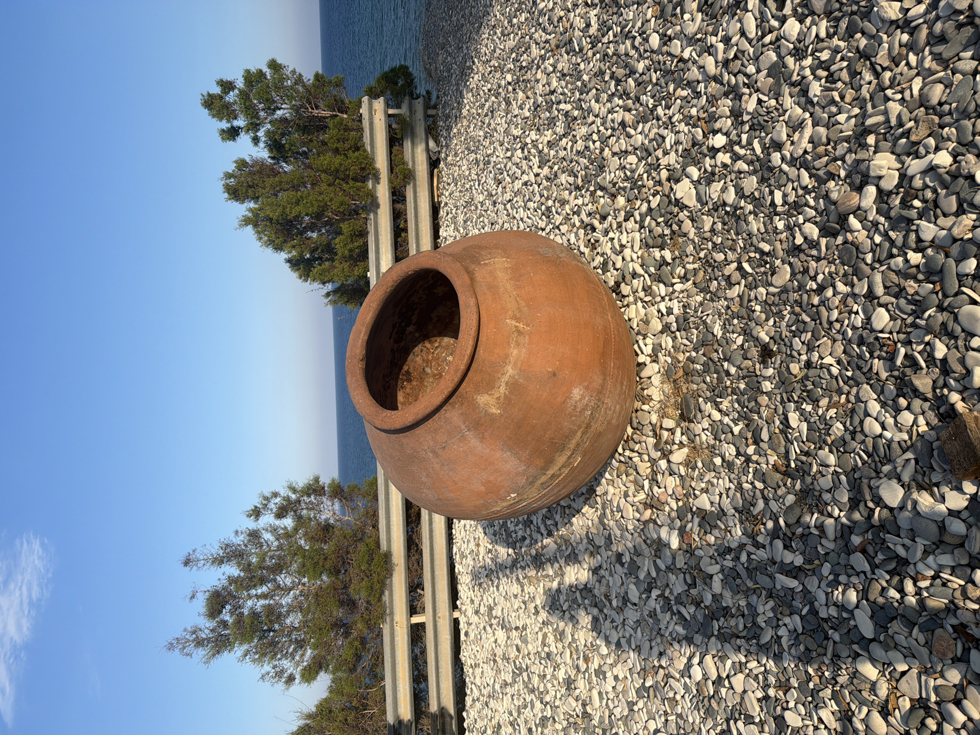 A large, reddish-brown terracotta pot rests on a bed of smooth, grey and white pebbles. The pot is round and wide, with a circular opening at the top. Its surface is textured and shows signs of age. In the background, a metal guardrail runs horizontally, separating the pebble area from a body of water, likely the sea, which is a deep blue. Behind the guardrail, several green, conical trees stand, their branches reaching upwards. The sky above is a clear, pale blue. The lighting suggests it is either early morning or late afternoon, casting long shadows from the pot onto the pebbles. The scene is located in Pervolia, Cyprus.