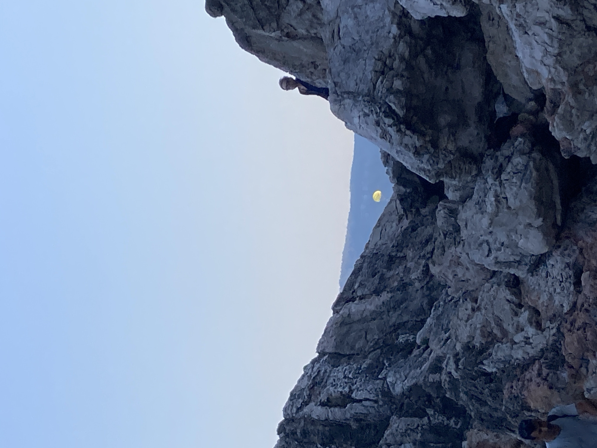 The image depicts a rocky, mountainous landscape with a clear blue sky. In the mid-ground, a person with short, dark hair, wearing a dark garment, is positioned on top of a large rock formation, looking to their left. To the left of this individual, further in the distance and partially obscured by rocks, a yellow, circular object is visible against the lighter sky, possibly the moon or a celestial body. In the foreground, to the left, a person wearing a light-colored t-shirt and a backpack is walking. The terrain is characterized by rugged, gray rocks and sparse vegetation. The overall setting suggests an outdoor, natural environment. It is not possible to determine the specific location within Kemer, Turkey from the visual information provided.