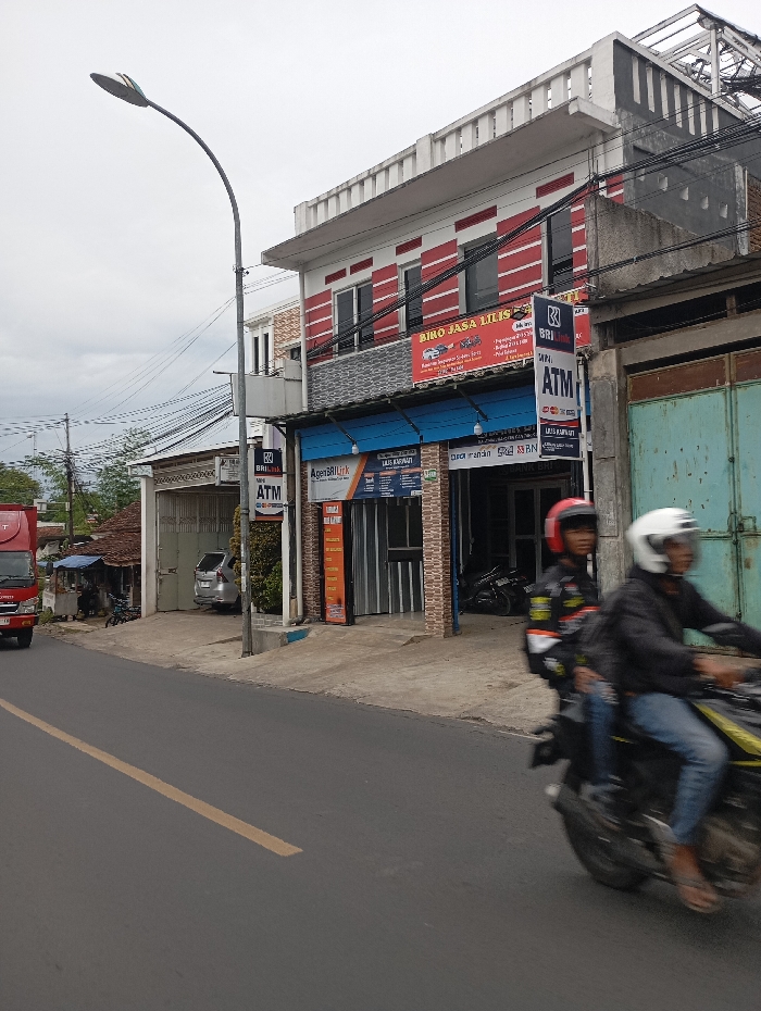 The image shows a street in Kecamatan Kutawaringin, Indonesia, with a two-story building on the right side of the frame. There is a sign on the building that reads "BIRO JASA Lilis," which is a local business. There is an ATM machine and a motorcycle parked in front of the building. A man is sitting on a motorcycle, wearing a helmet and looking ahead. In the background, there is a truck parked on the street. The sky is cloudy and the street is paved with asphalt. There is a yellow dividing line on the road. The image was taken on a street with power lines running overhead. The street is flanked by a sidewalk on the right side of the image. There is a streetlight positioned above the sidewalk. There is a small, white car parked on the left side of the frame. The image is shot from street level.