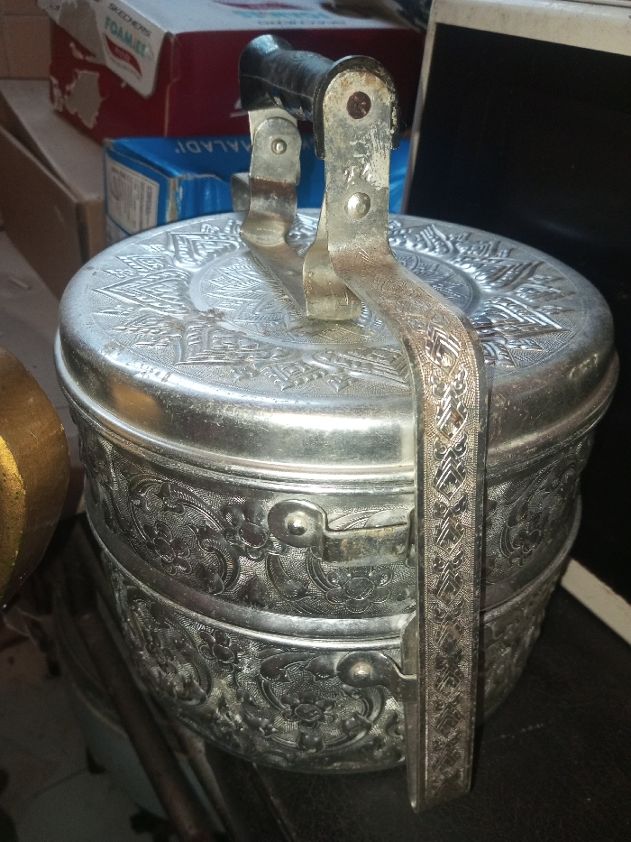 A tiered, ornate silver tiffin carrier with a black handle sits prominently. The tiffin has detailed floral and geometric engravings. Oxidization is visible, giving it an antique appearance. The carrier is secured by a silver handle. Partially visible are cardboard boxes and a tablet. The overall scene suggests a storage area or a cluttered household setting.